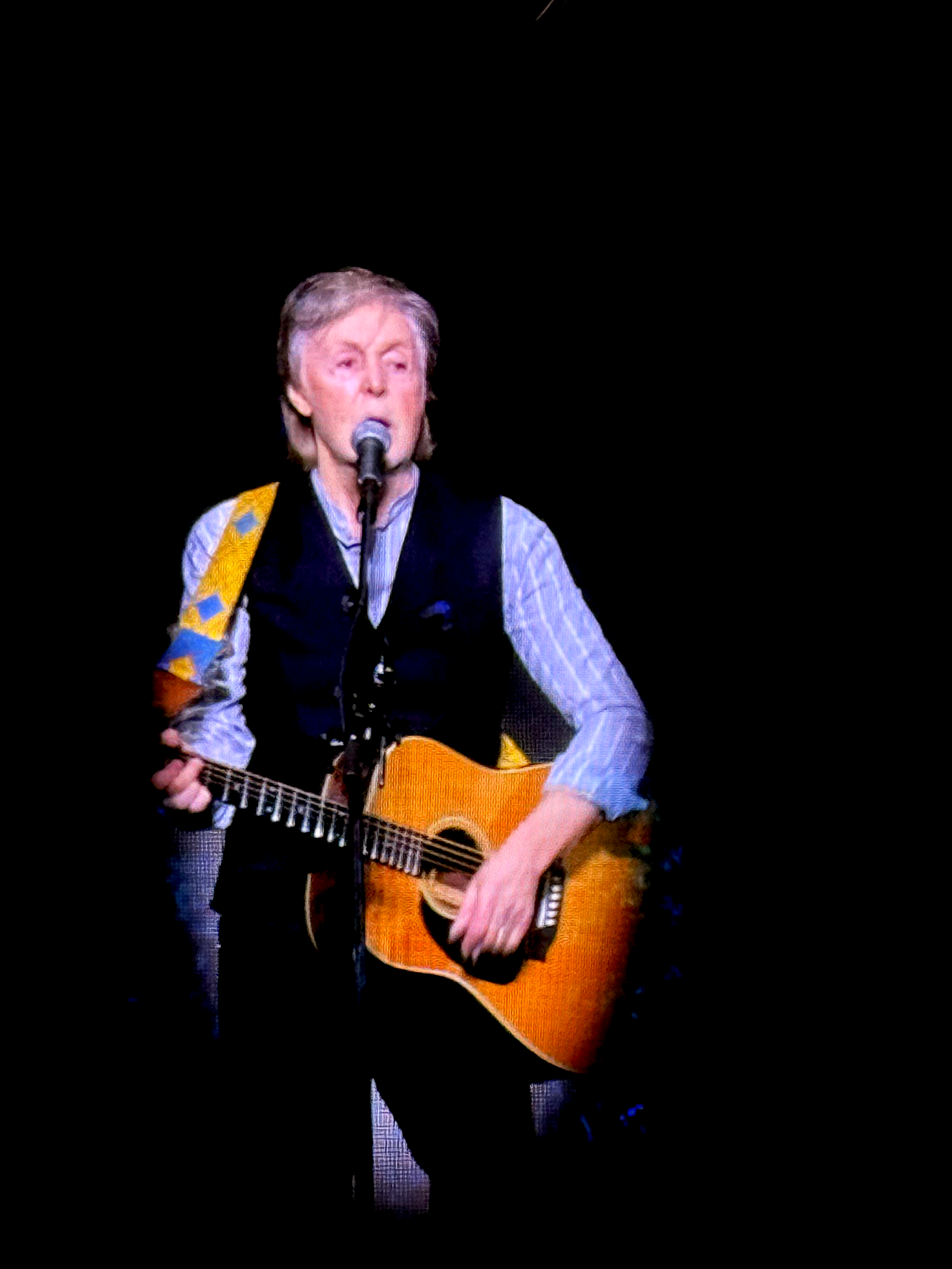 An elderly woman with gray hair singing into a microphone while playing an acoustic guitar on a dark stage.