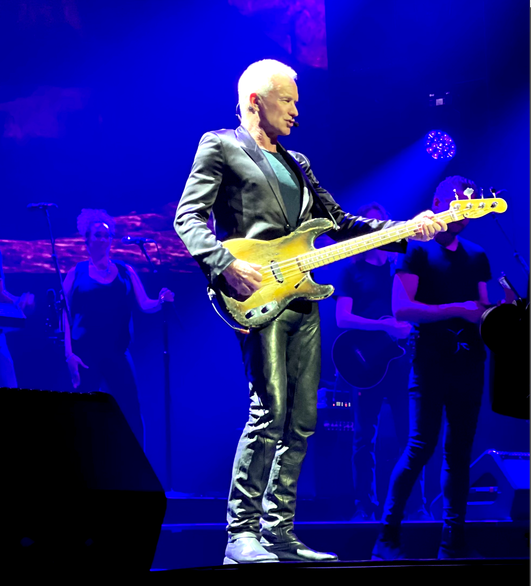 Sting on stage at The Palladium April 2022., playing an electric bass guitar with backup singers and other musicians in the background, illuminated by blue stage lighting.
