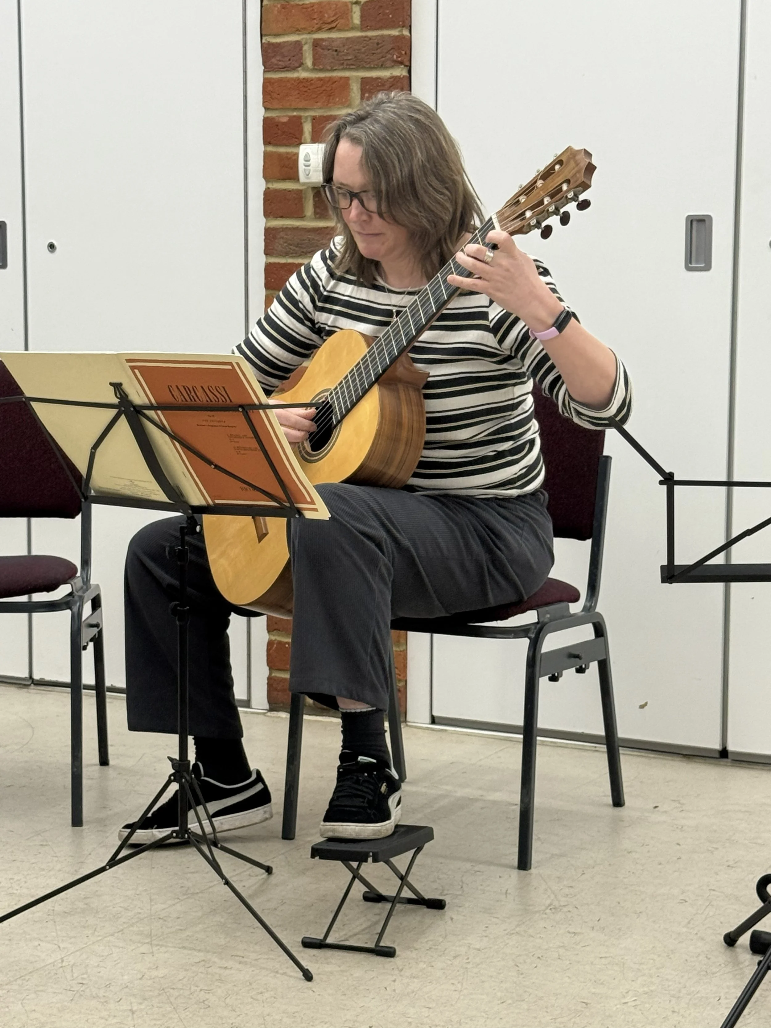 Person playing an acoustic guitar while sitting on a chair, reading sheet music, in an indoor setting with brick and white wall background.