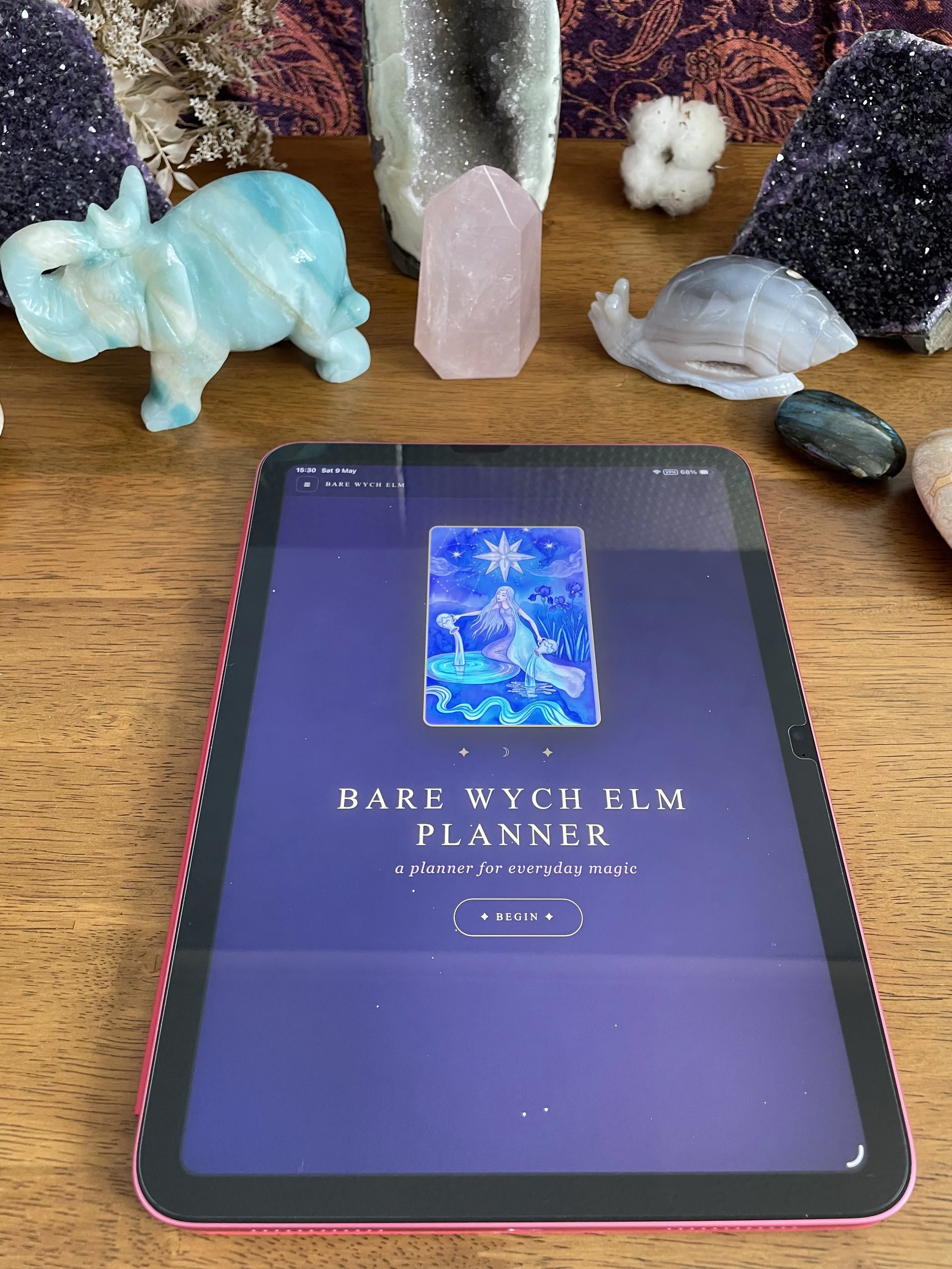 An electronic tablet displaying the cover of "Bare Wych Elm Planner," a digital planner for everyday magic, positioned on a wooden surface surrounded by various crystals and stones, including amethyst, rose quartz, and labradorite.