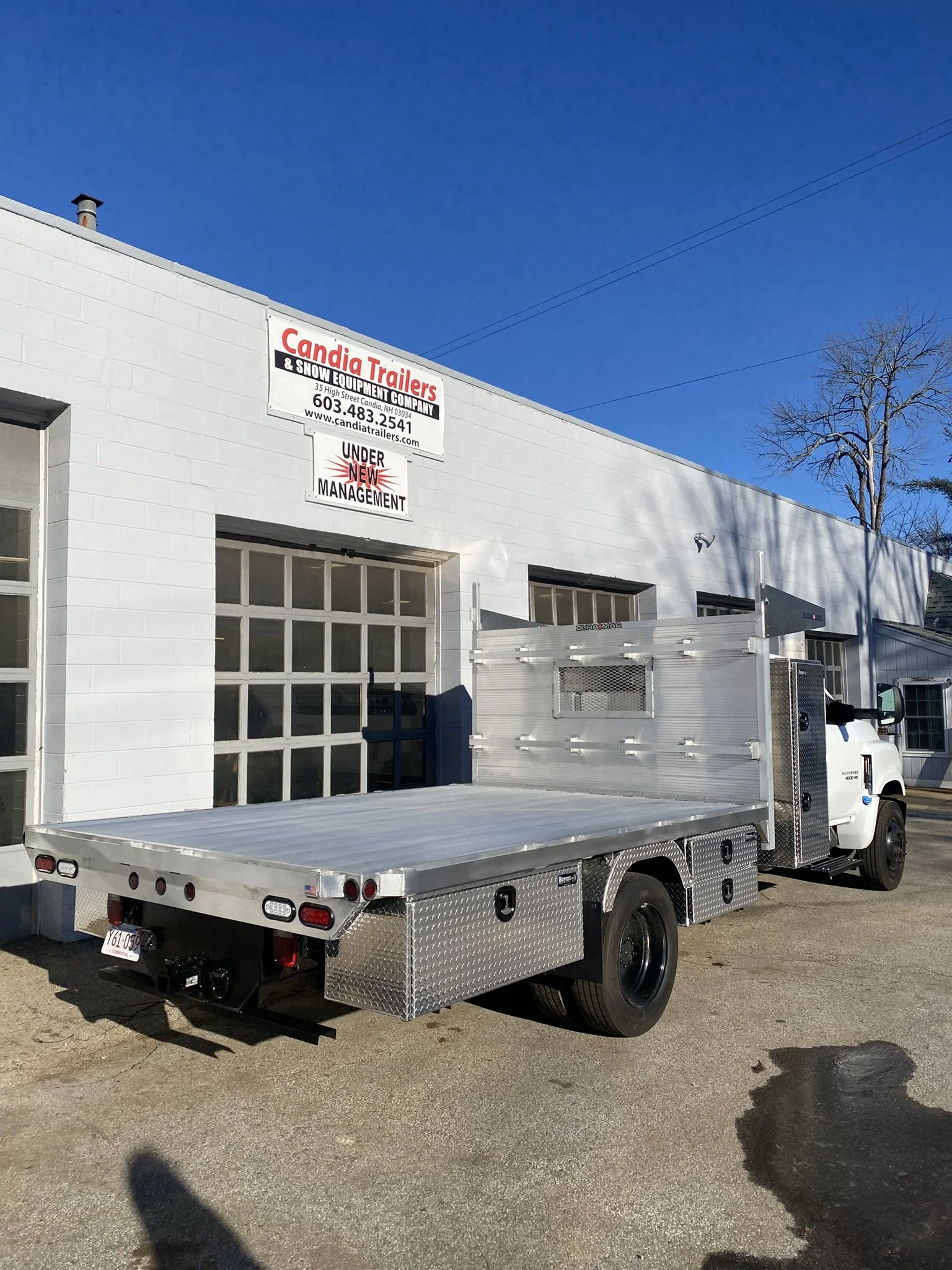Aluminum flatbed utility truck parked outside a white building with signs for Candia Trailers and Snow Equipment Company, under a clear blue sky.