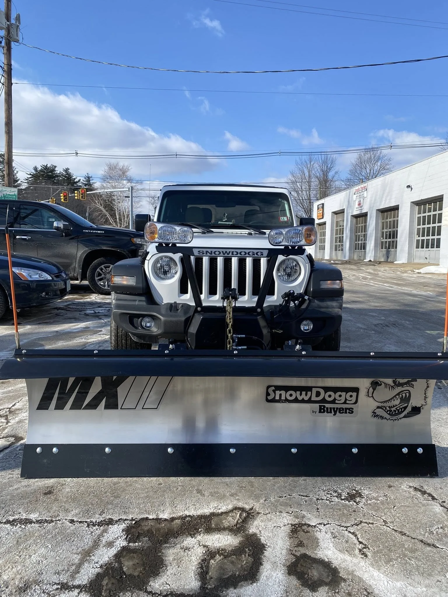Front view of a white Jeep with a snow plow attachment at the front, parked on a snowy and icy lot with other vehicles and a white building in the background.