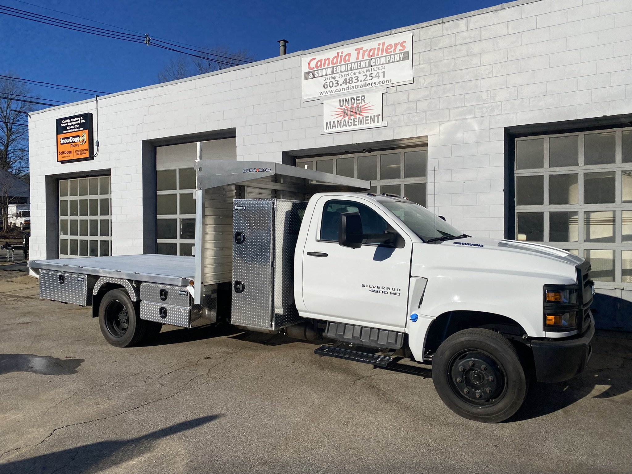 White utility truck with aluminum flatbed and storage compartments parked in front of a white building with signs, including 'Candia Trailers' and 'Snow Equipment' storefronts.