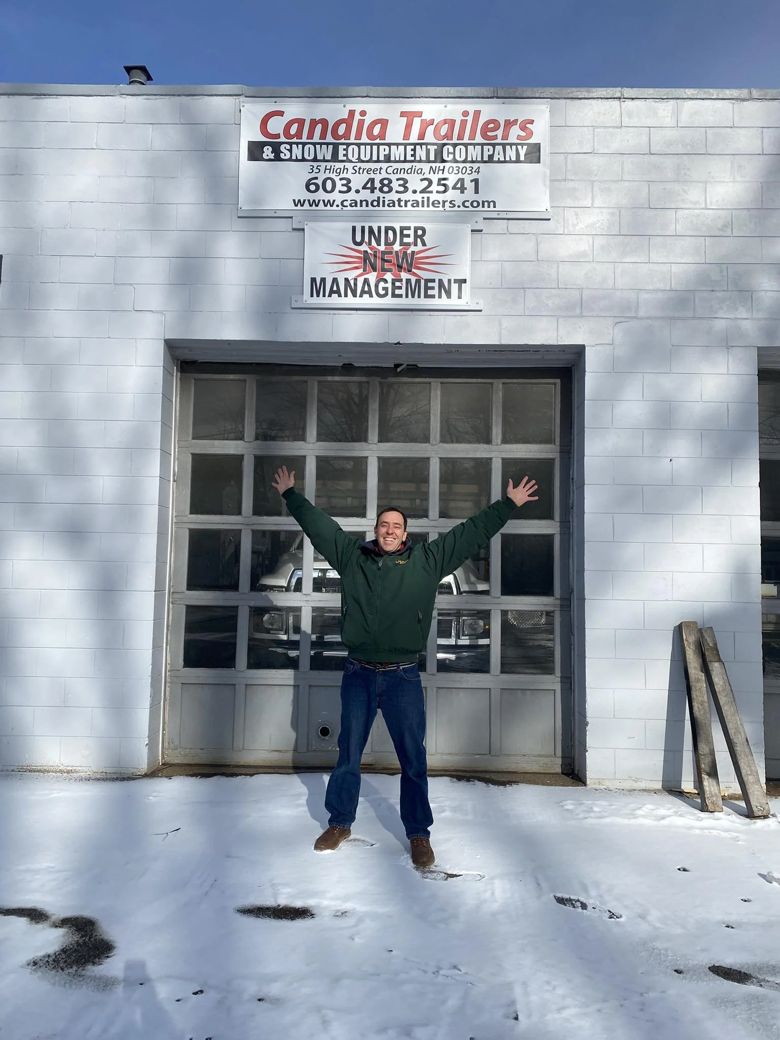 Man standing outside Candia Trailers & Snow Equipment Company in front of the garage door with arms raised in celebration, snow on the ground, clear sky above.