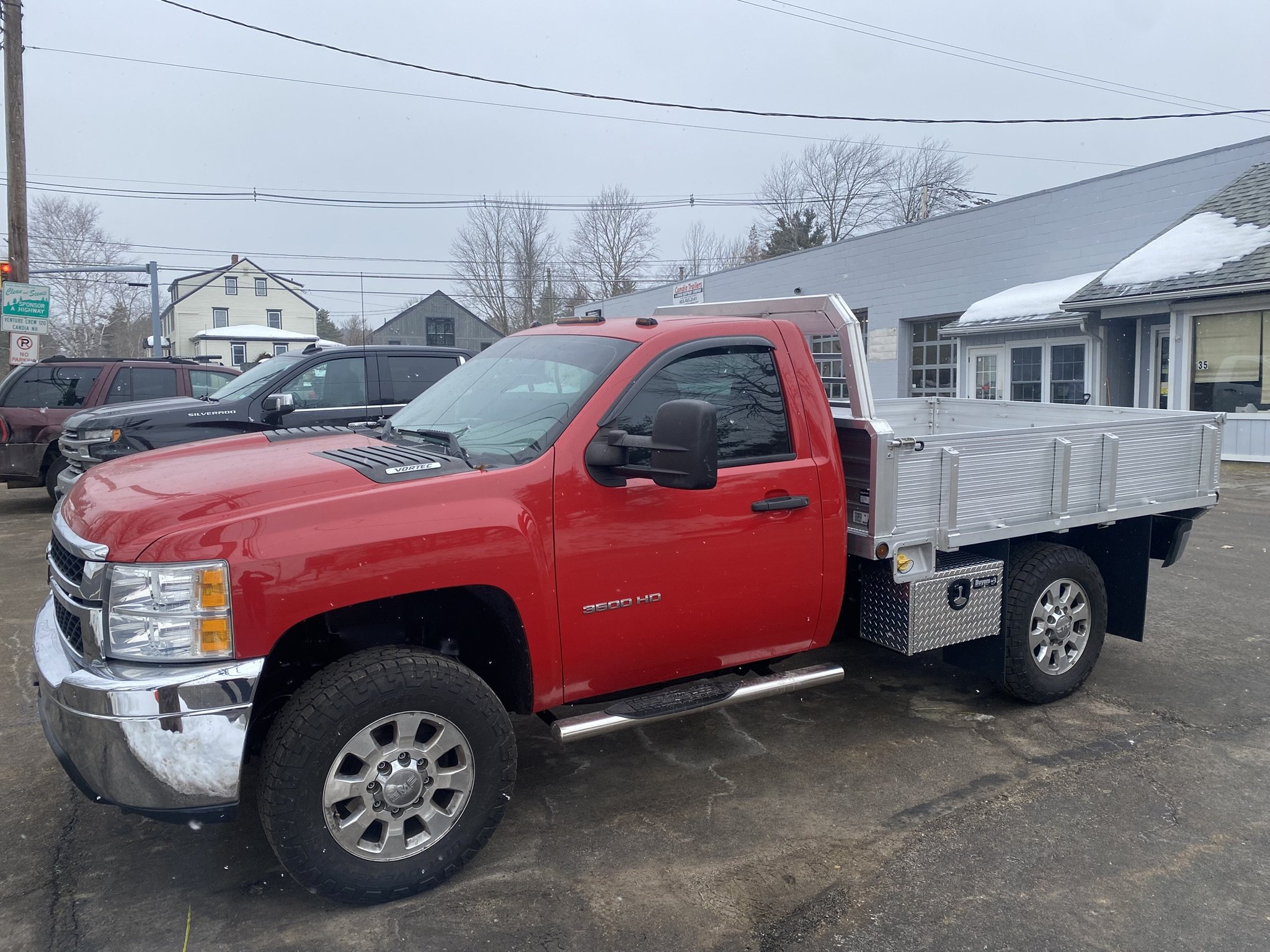 A red flatbed utility truck parked in a lot with other vehicles and buildings in the background, on a cloudy winter day.