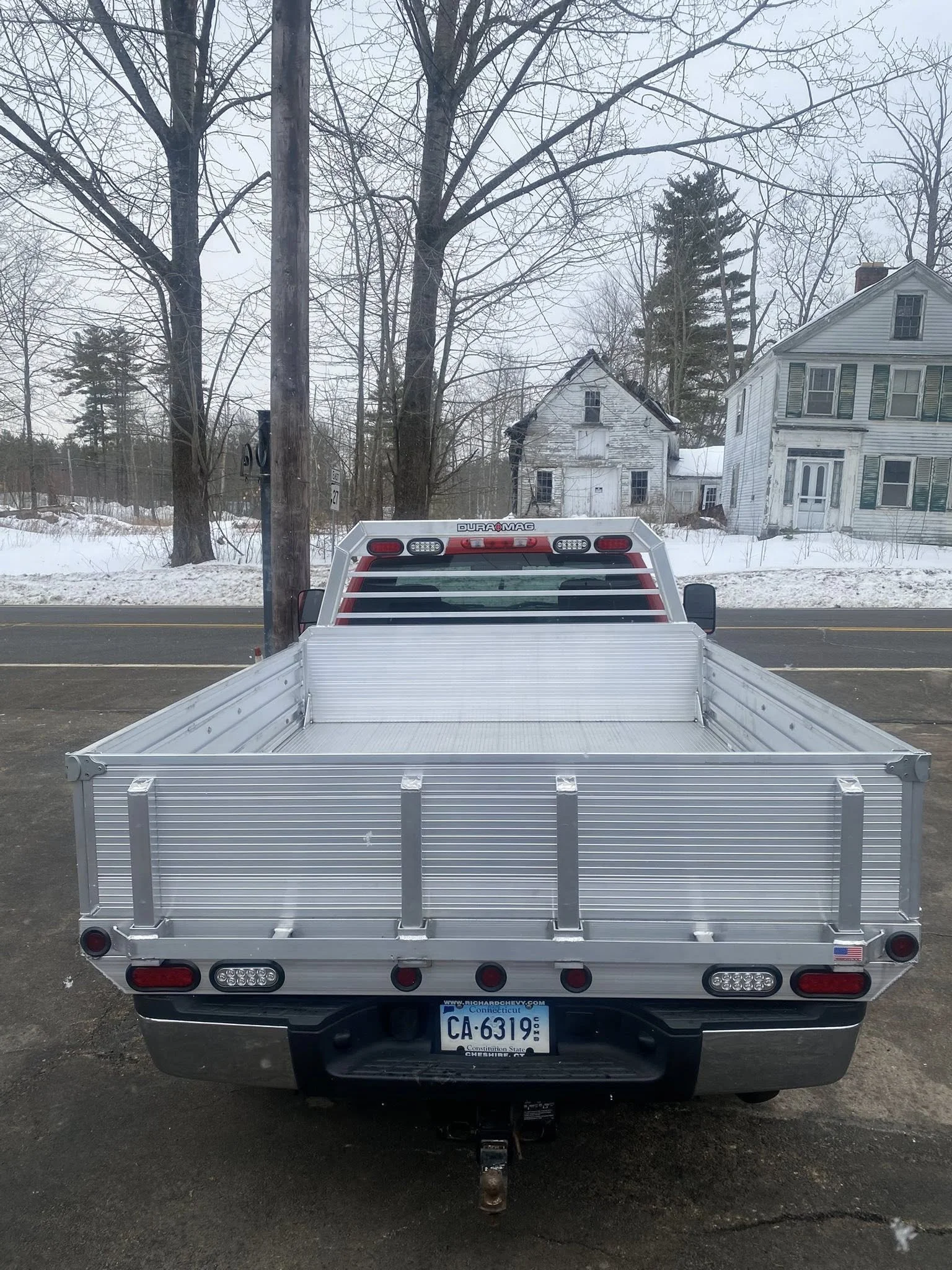 Front view of a silver flatbed truck with a Connecticut license plate, parked on the side of a road with snow. Houses and trees are visible in the background.