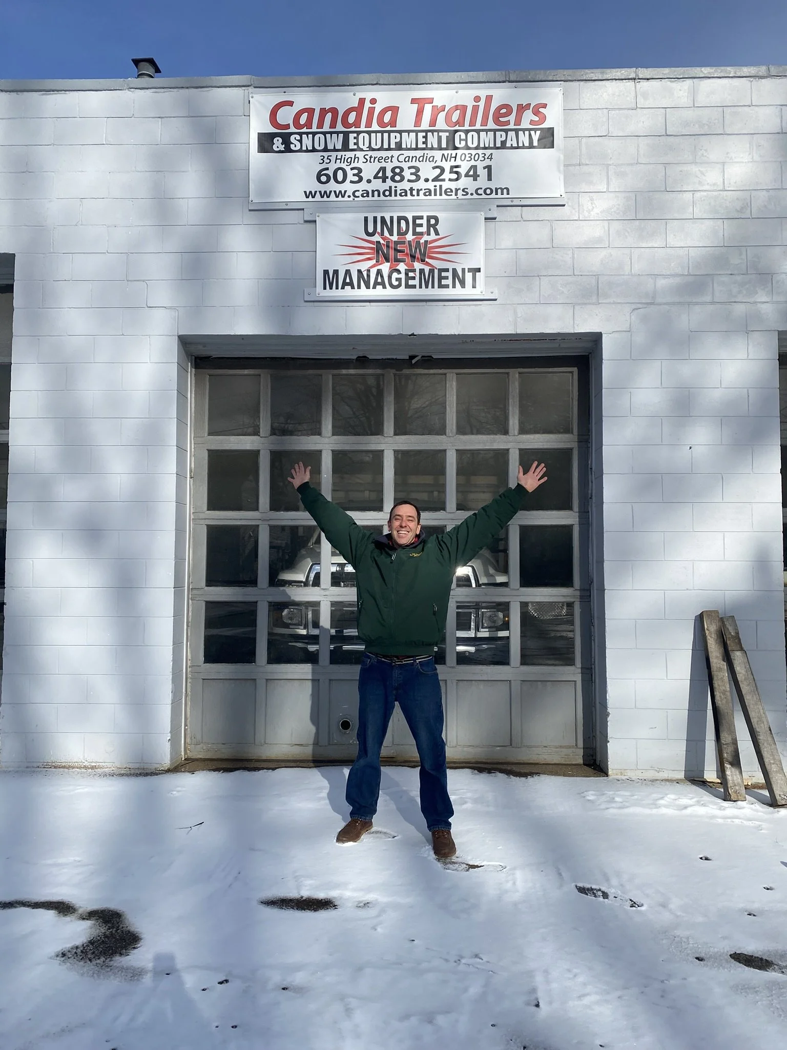 Man standing outside an industrial building, smiling with arms raised, snow on the ground, signs above reading 'Candia Trailers & Snow Equipment Company' and 'Under New Management'.