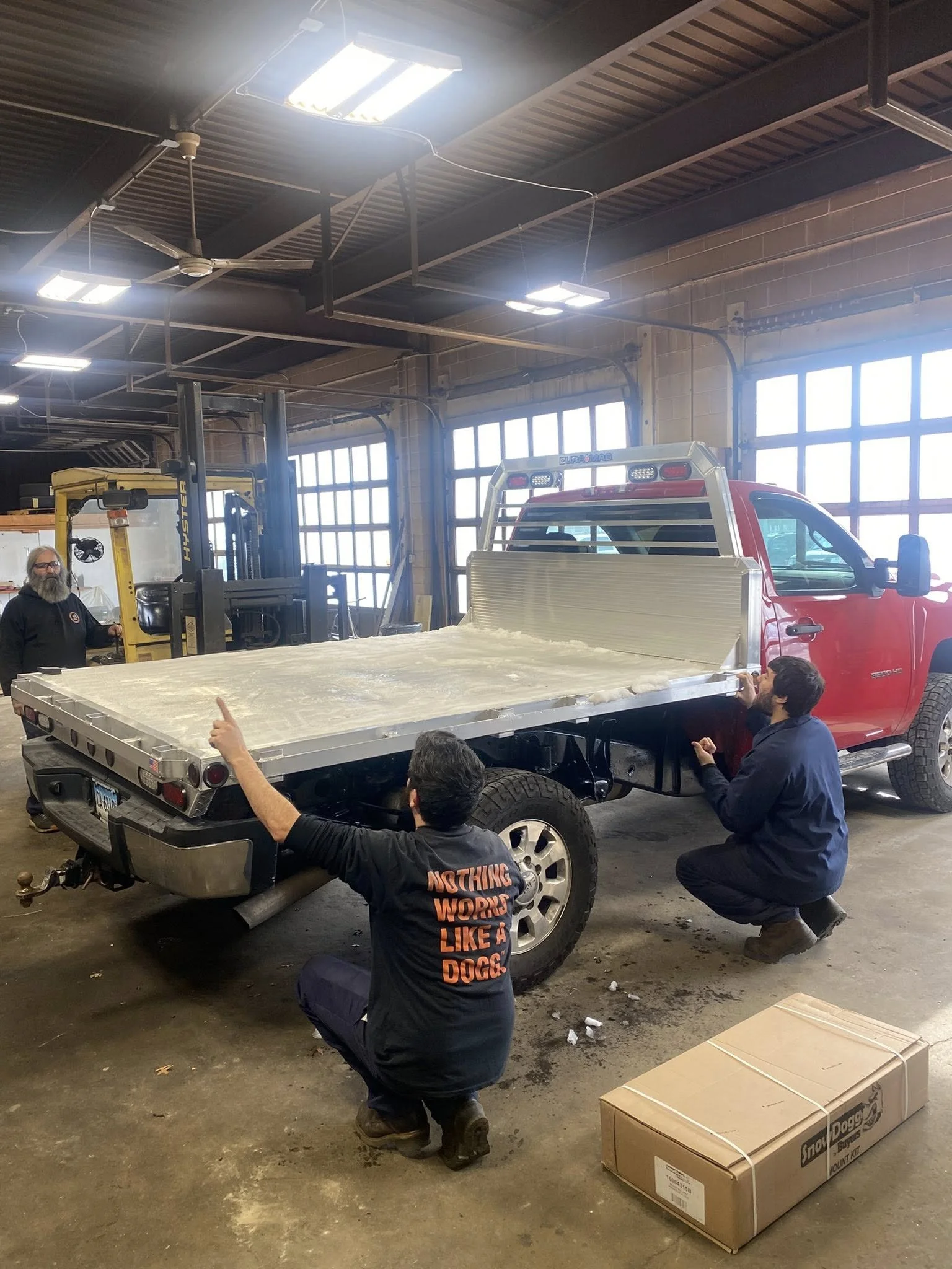 Three people working on a red utility truck inside a workshop, with two kneeling and one standing, near the truck's flatbed, which is in the process of being assembled.