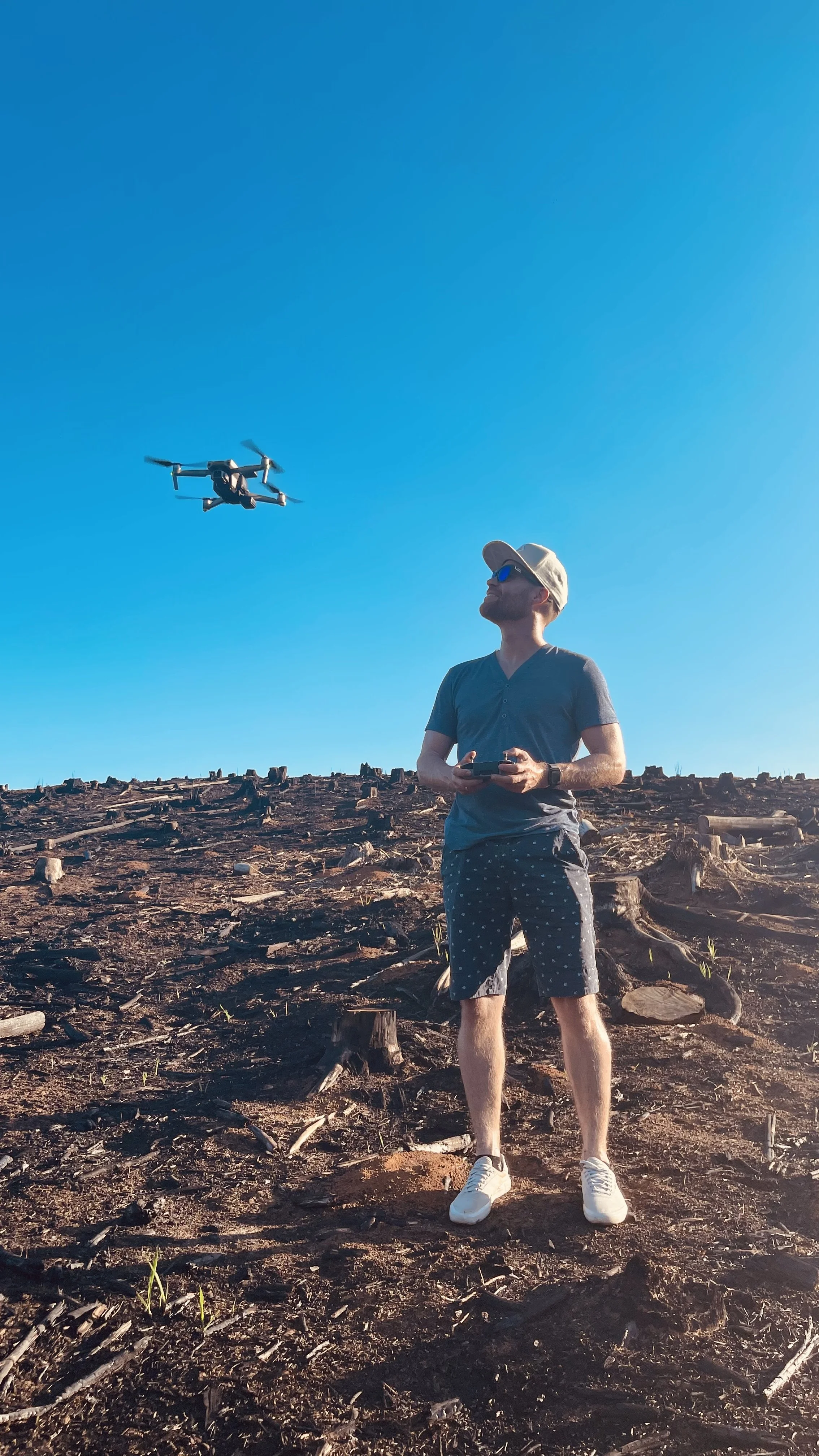 Erik van der Ven wearing a hat and sunglasses is standing on a field of cut trees, holding a remote control, with a drone flying in the blue sky above him.