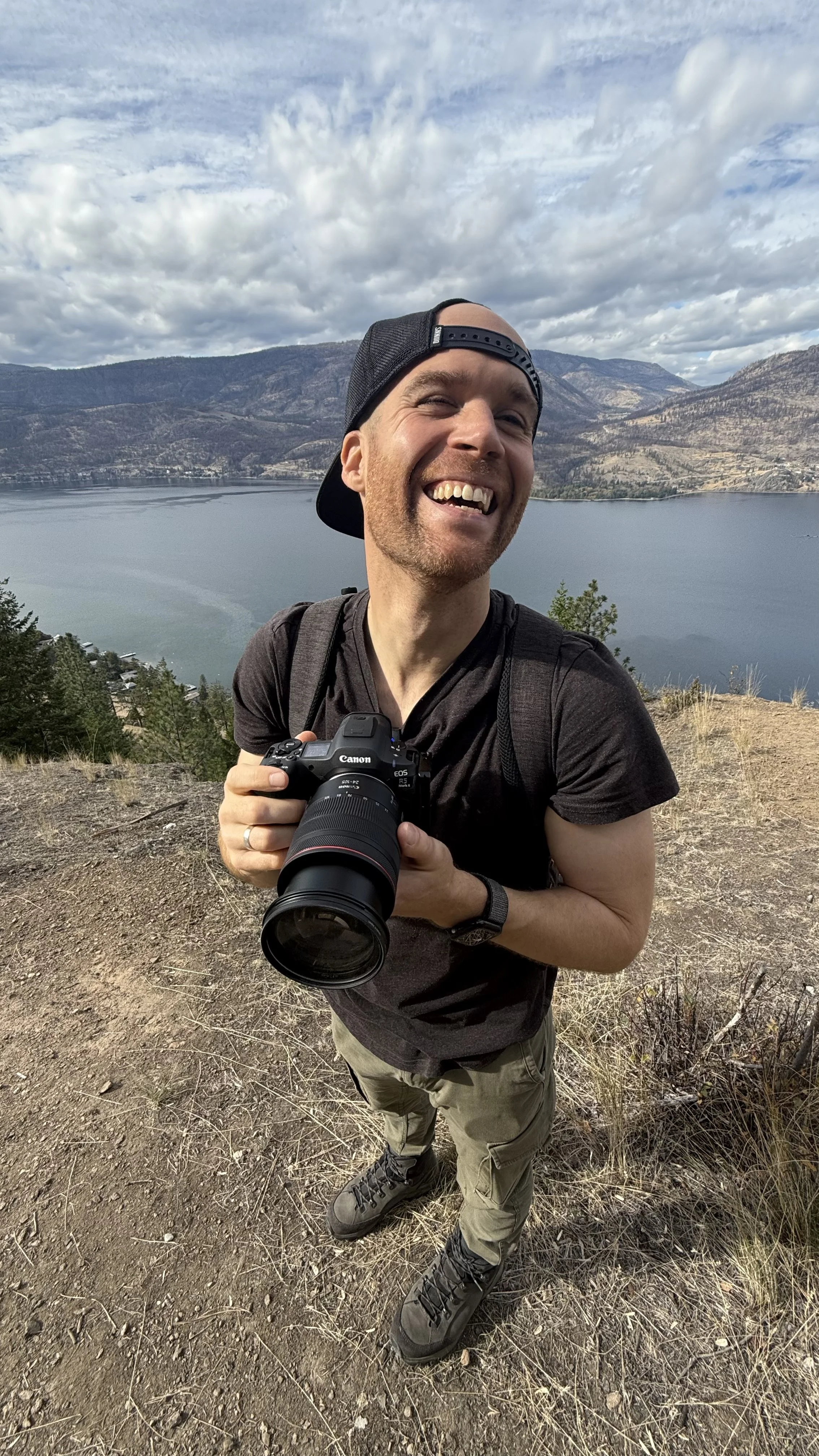 Erik van der Ven with a Canon camera around his neck smiling and looking at the camera, outdoors with mountains, lake, and partly cloudy sky in the background.