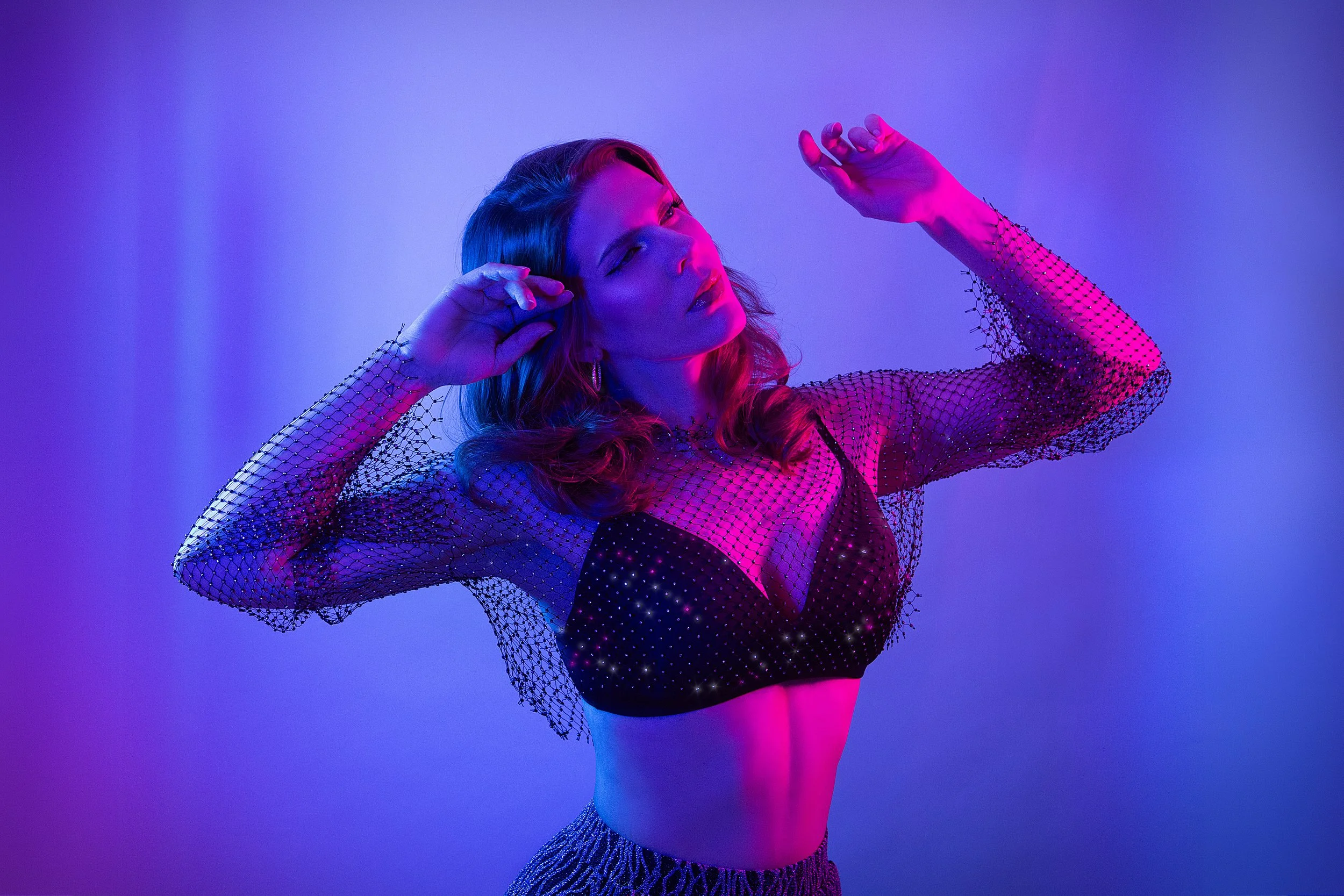 A woman with wavy hair posing against a purple and pink gradient background, wearing a sheer dotted top with a black bra and a ruffled skirt.