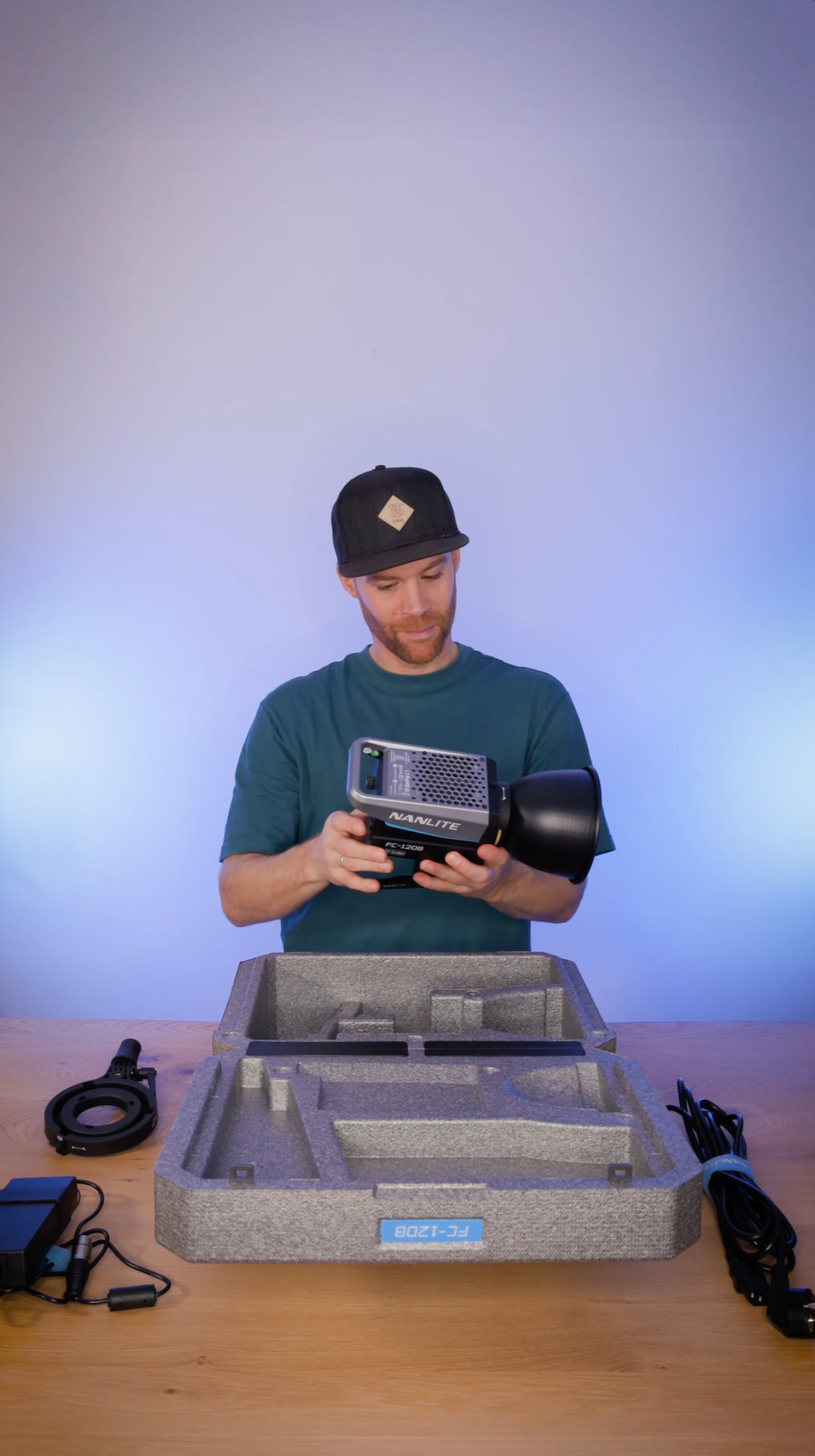 Erik van der Ven with a beard and a black cap is holding a photography lighting strobe in a foam-lined box on a wooden table, with camera accessories and cables around.