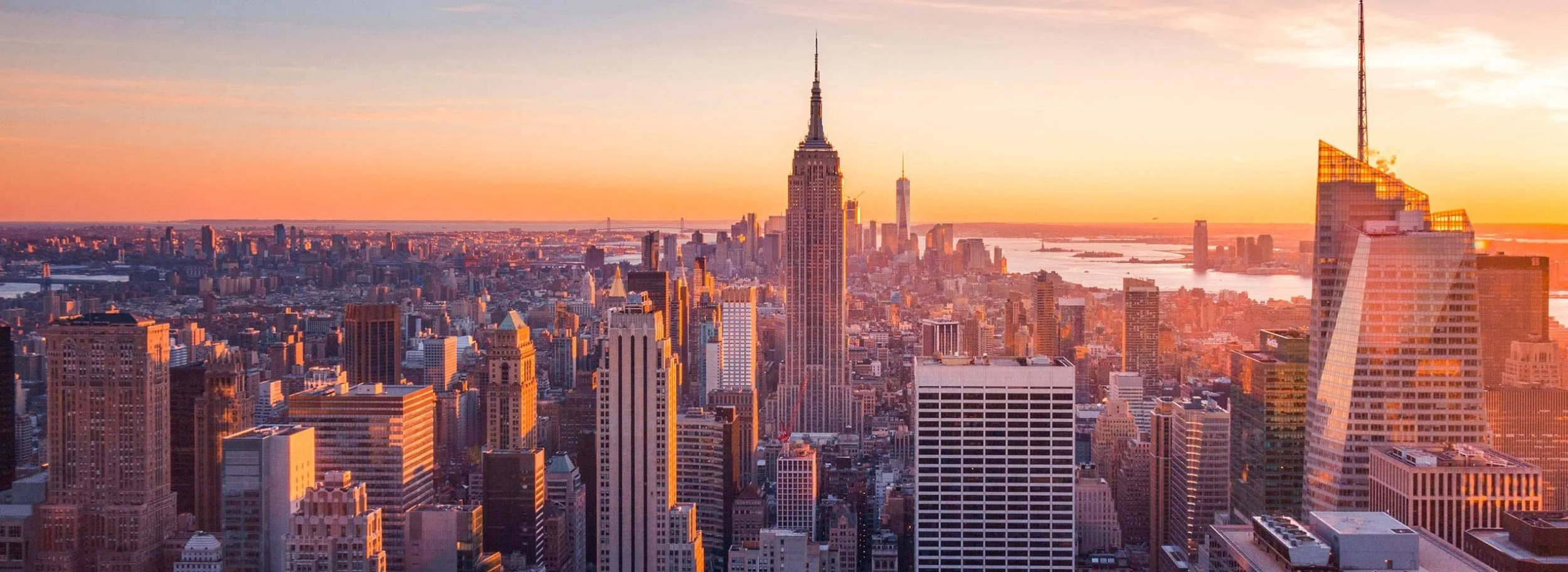 Sunset over New York City skyline featuring the Empire State Building and surrounding skyscrapers with a harbor in the background.