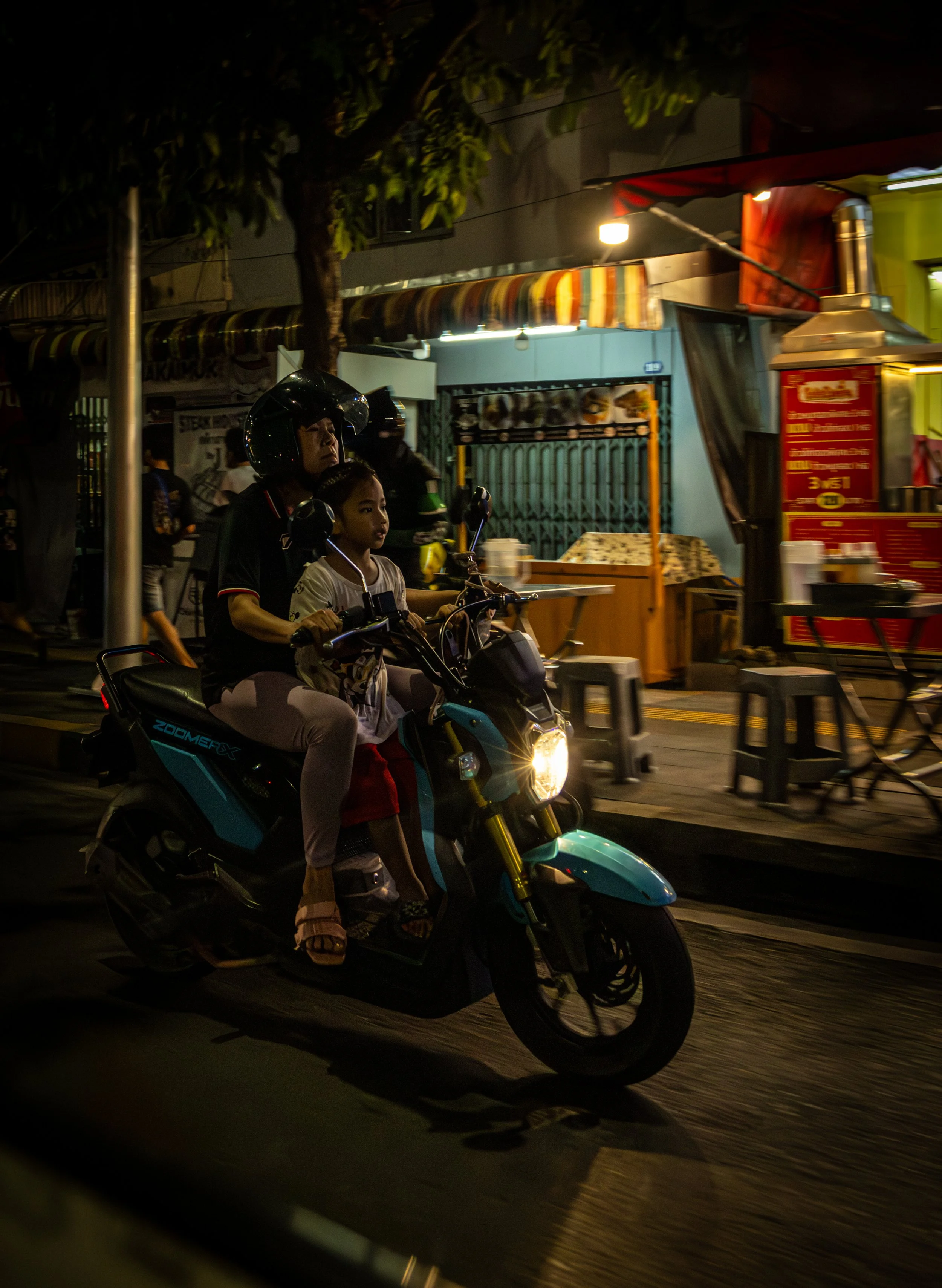 A person and a young girl riding a scooter at night on a city street with market stalls and signs illuminated in the background.