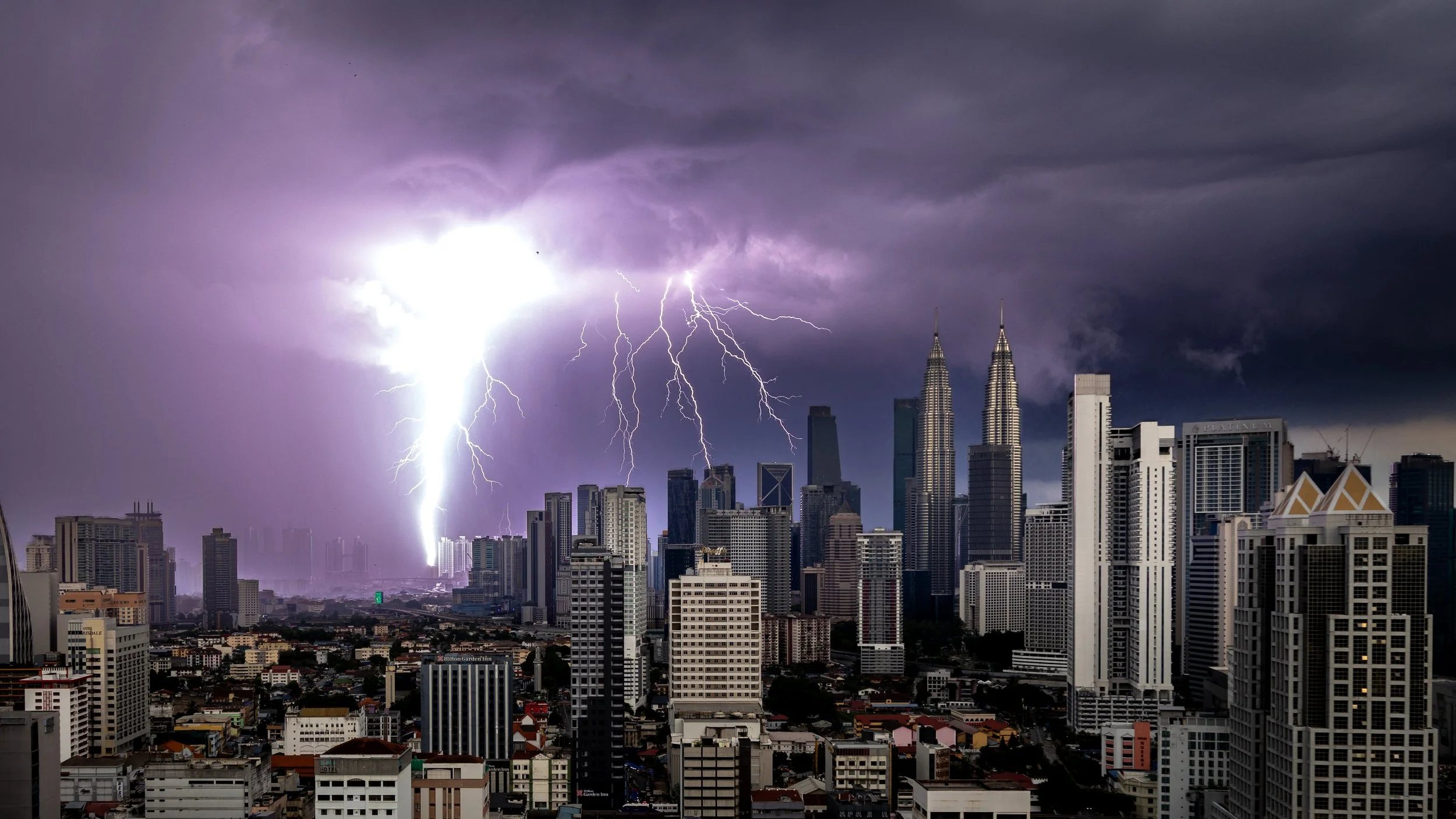 Lightning strikes over the Kuala Lumpur city skyline with tall buildings and dark storm clouds.