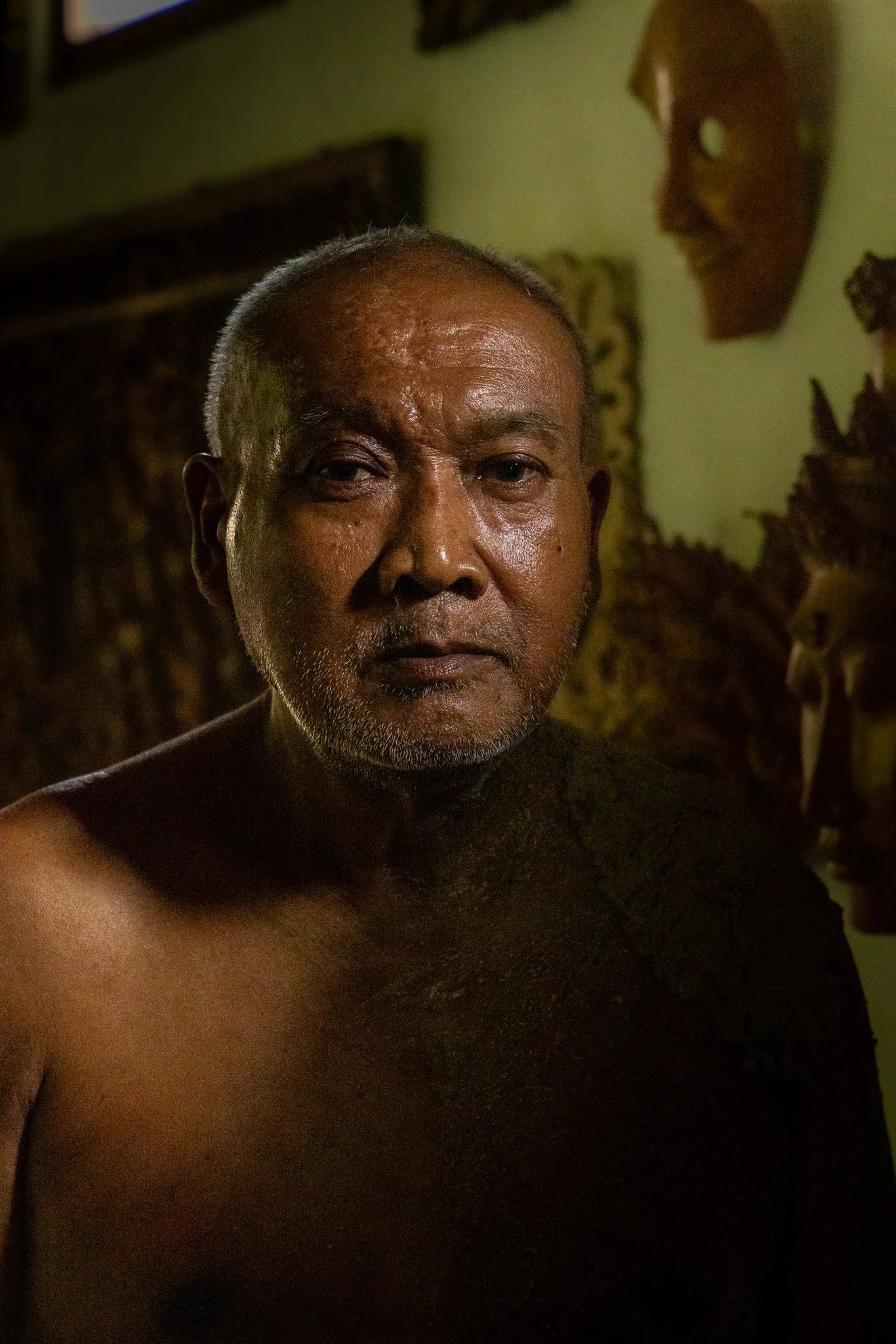 Close-up portrait of an elderly man with dark skin, short gray hair, and a calm expression. The background features traditional masks and wall decorations.