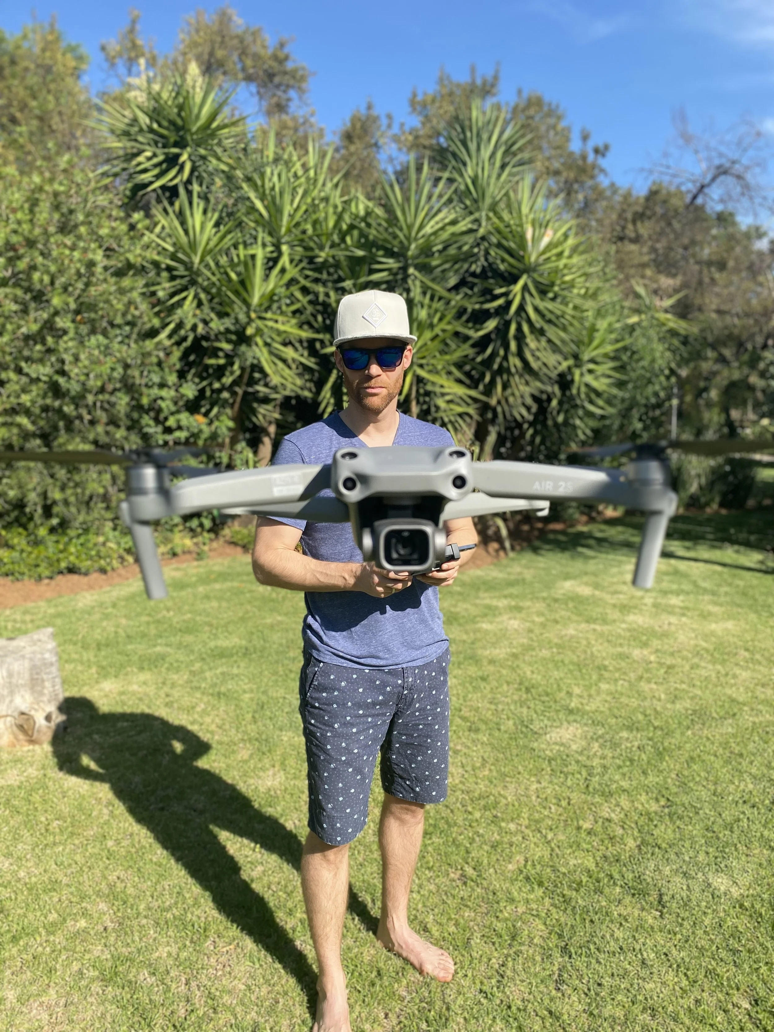 Erik van der Ven holding a drone in a grassy outdoor area with trees, wearing sunglasses, a white cap, a blue t-shirt, and patterned shorts.