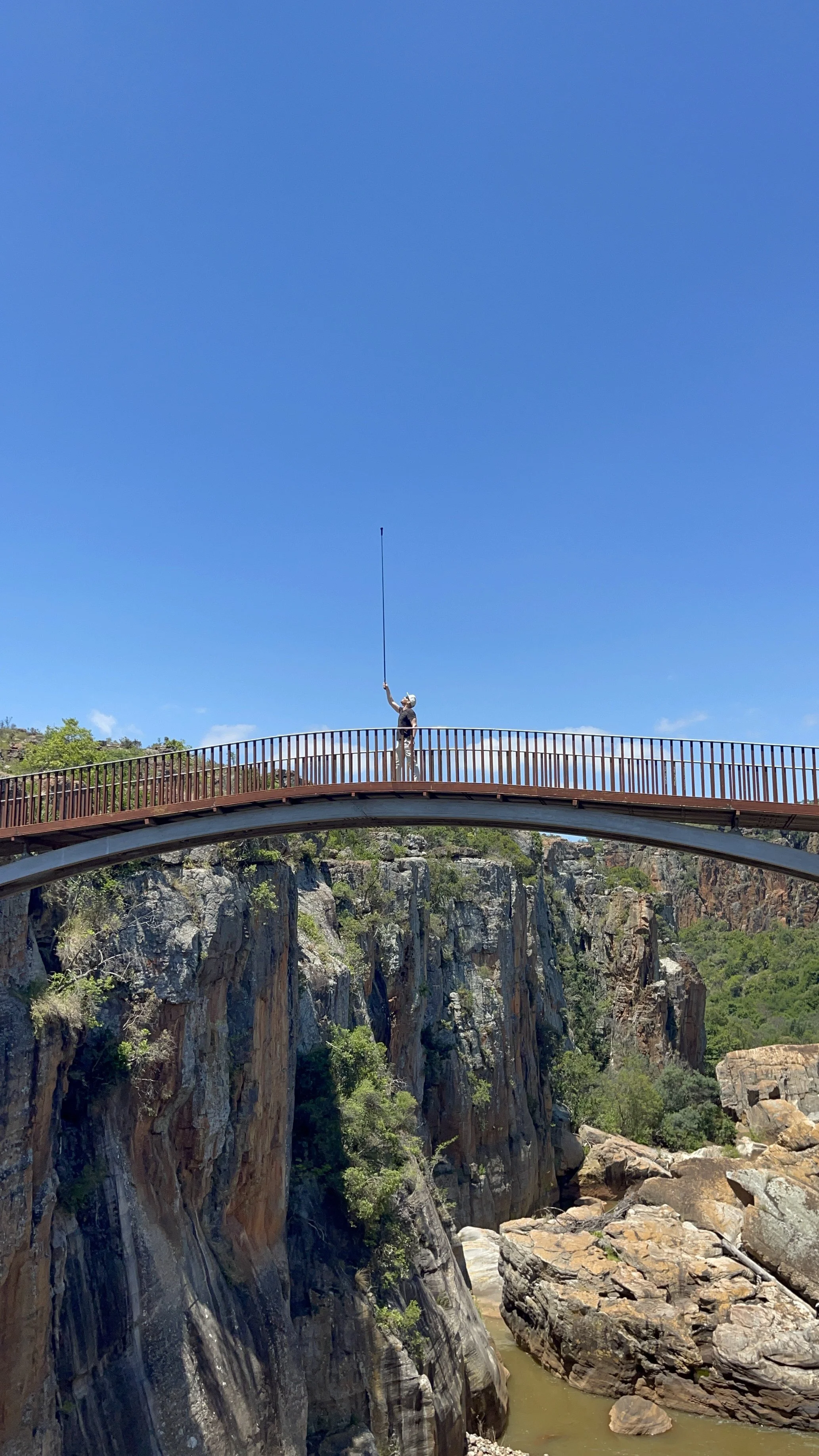 Erik van der Ven standing on a bridge over a canyon, with a long selfie stick under a clear blue sky.