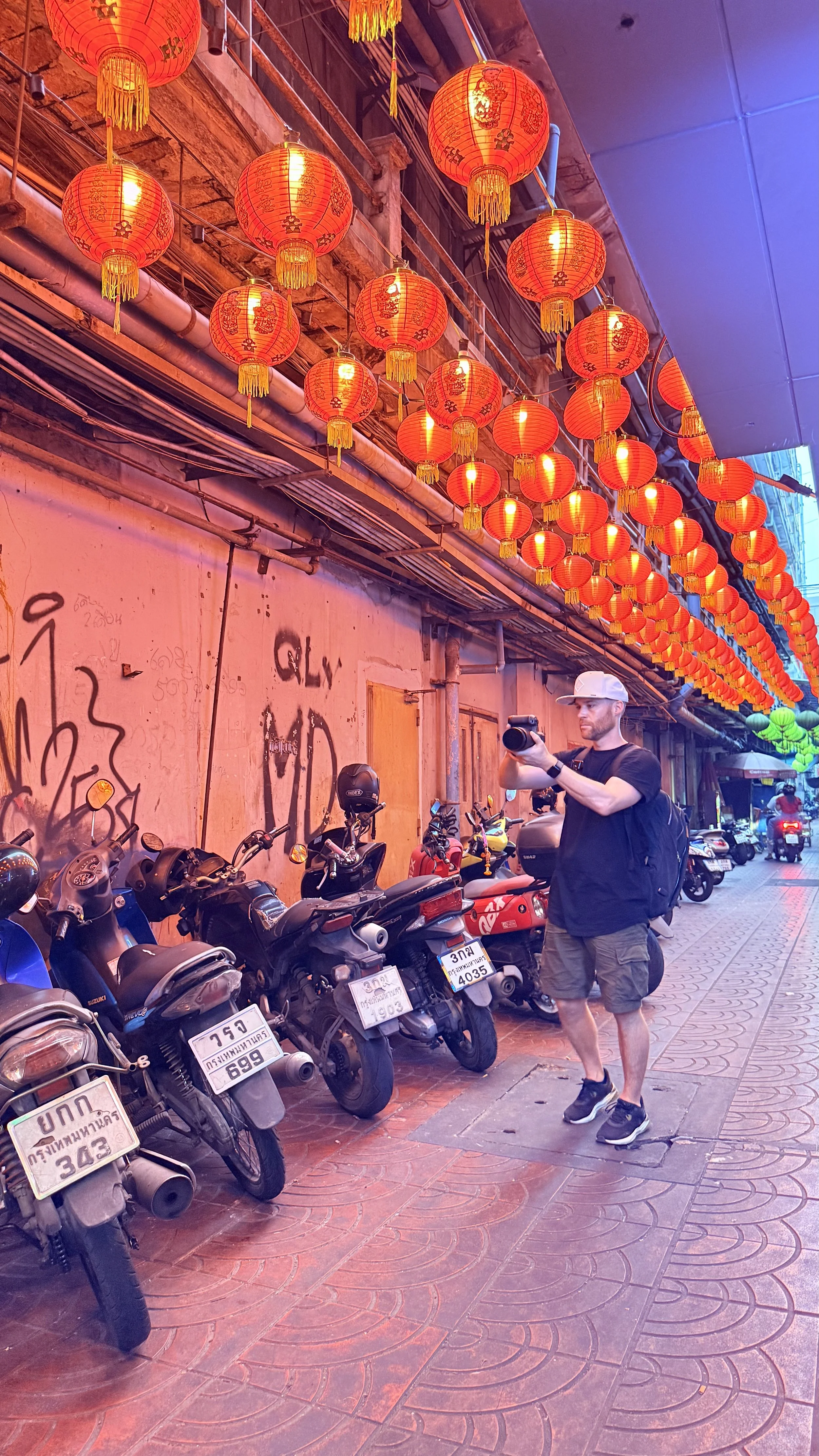 Street scene with decorative red Chinese lanterns hanging from the ceiling above a row of parked motorcycles. Erik van der Ven wearing a white cap, black t-shirt, and gray shorts is taking a photo. There is graffiti on the wall behind the motorcycles