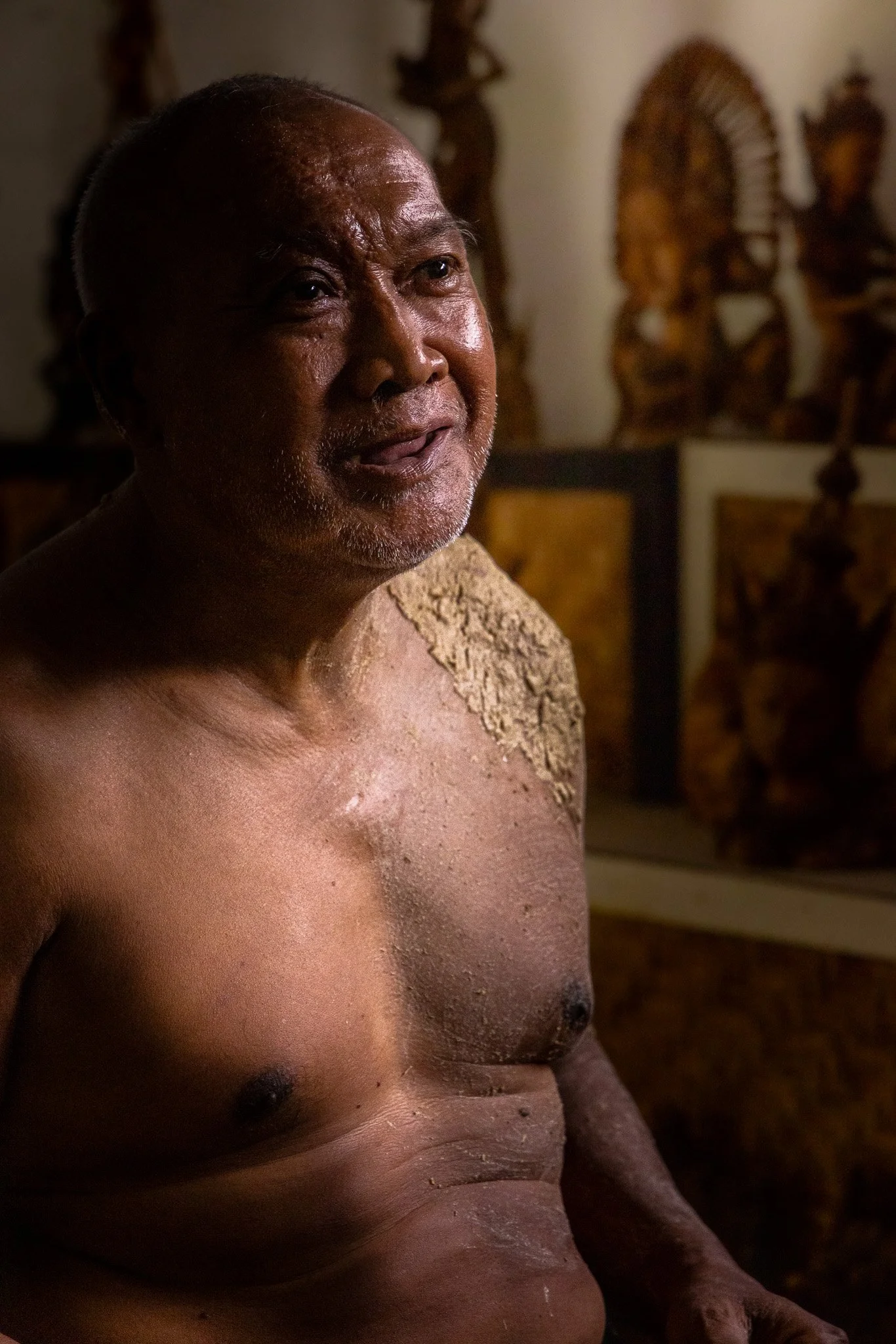 Portrait of a old man with a shaved head, smiling and looking to the side, in a dimly lit room with traditional wooden carvings in the background.