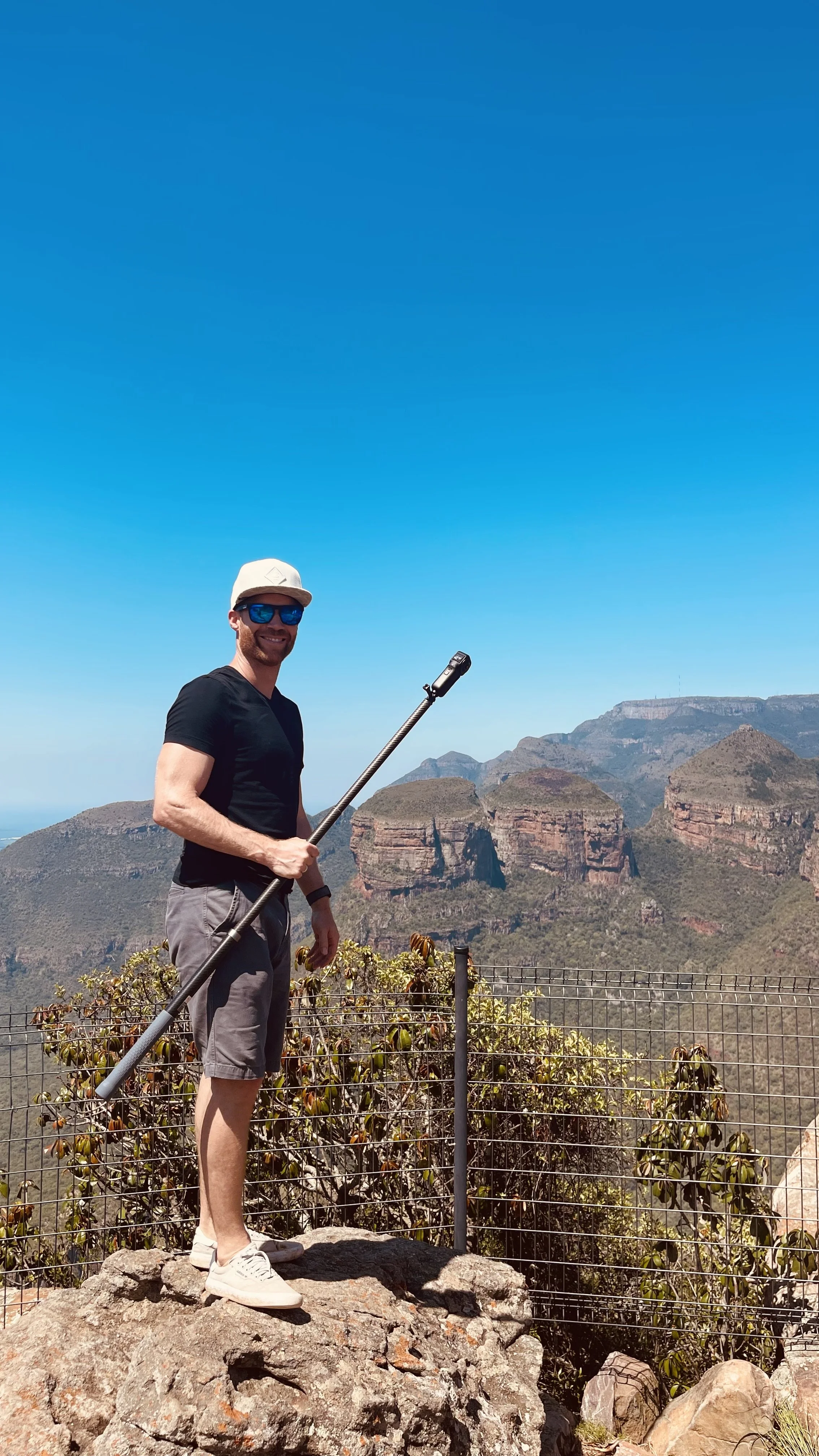 Erik van der Ven wearing a white cap, blue sunglasses, black t-shirt, and gray shorts holding a selfie stick while standing on a rocky area with a mountain range in the background under a clear blue sky.