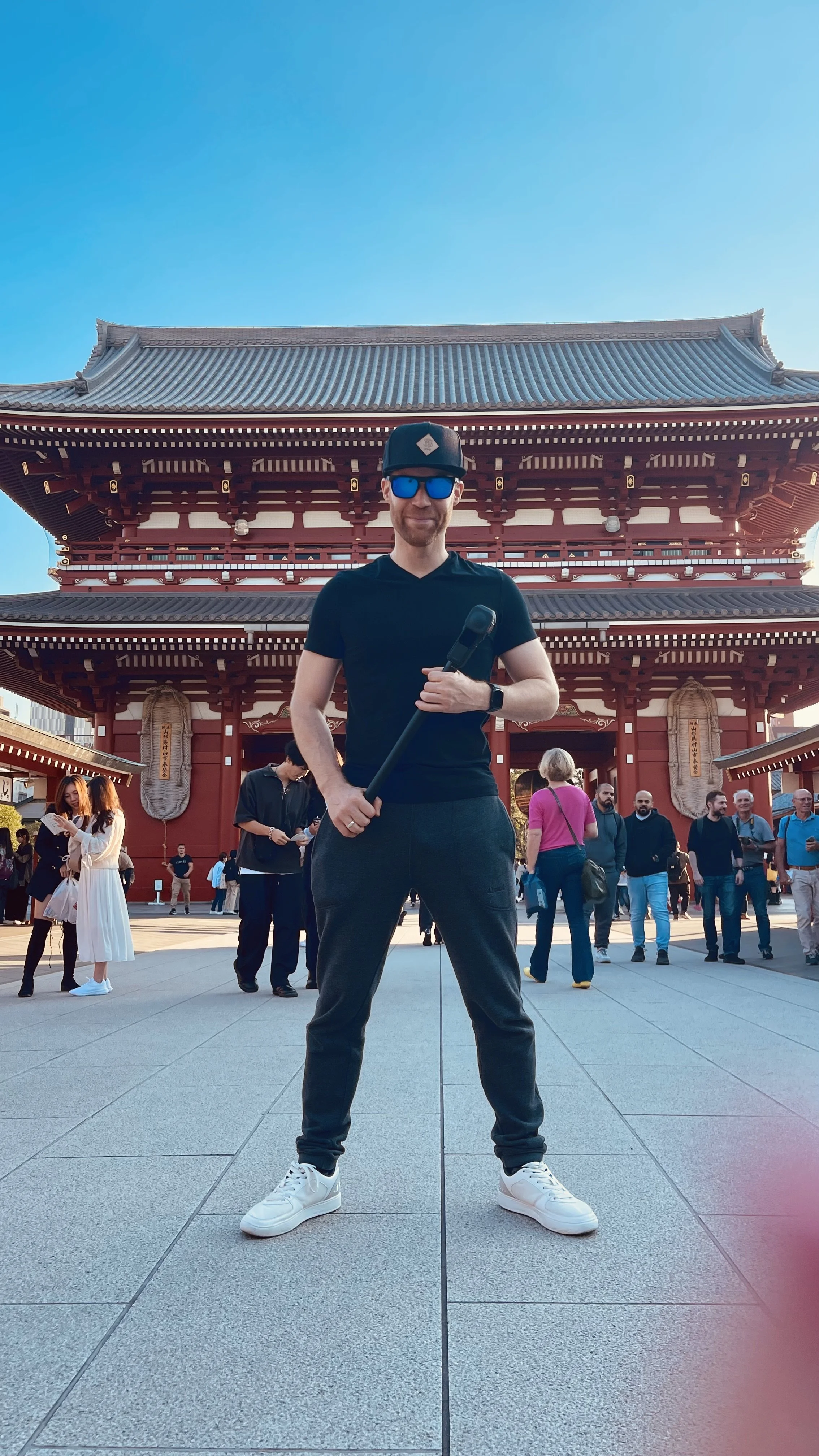 Erik van der Ven in black clothing and sunglasses in front of a traditional Japanese temple with a red facade and a gray tiled roof. Several people are seen in the background at the temple grounds, some talking and others taking photos.