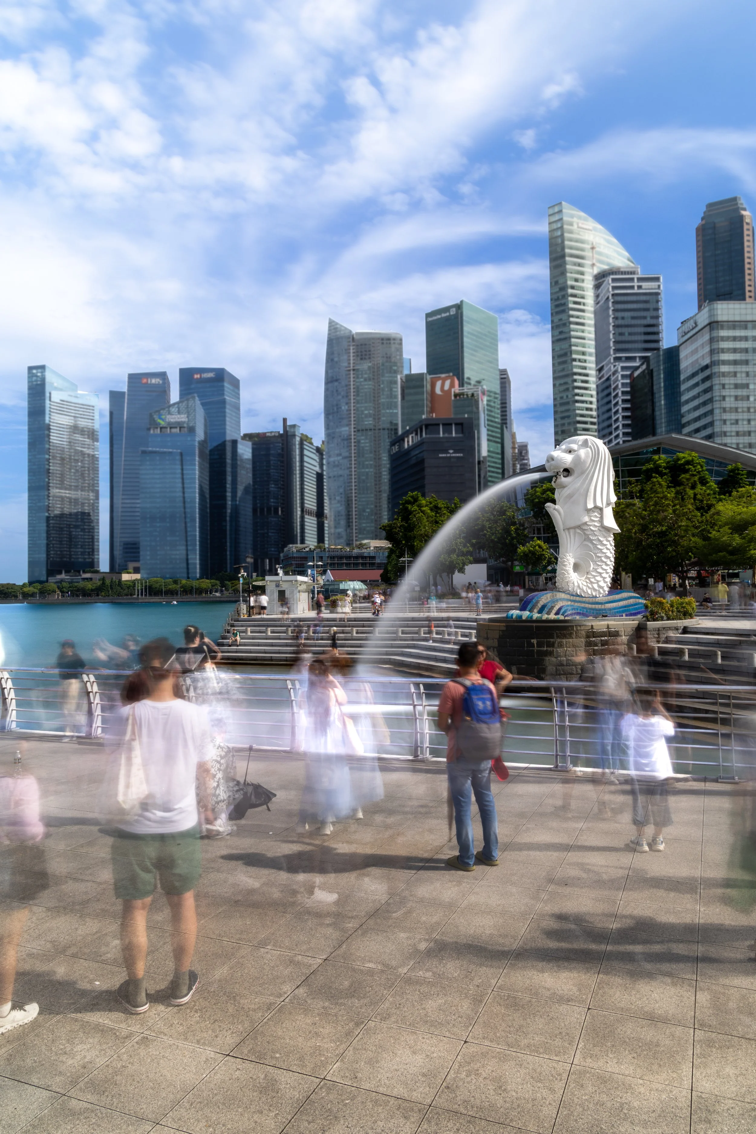 People walking on a waterfront promenade with the Merlion statue and a city skyline of tall skyscrapers in the background on a sunny day.
