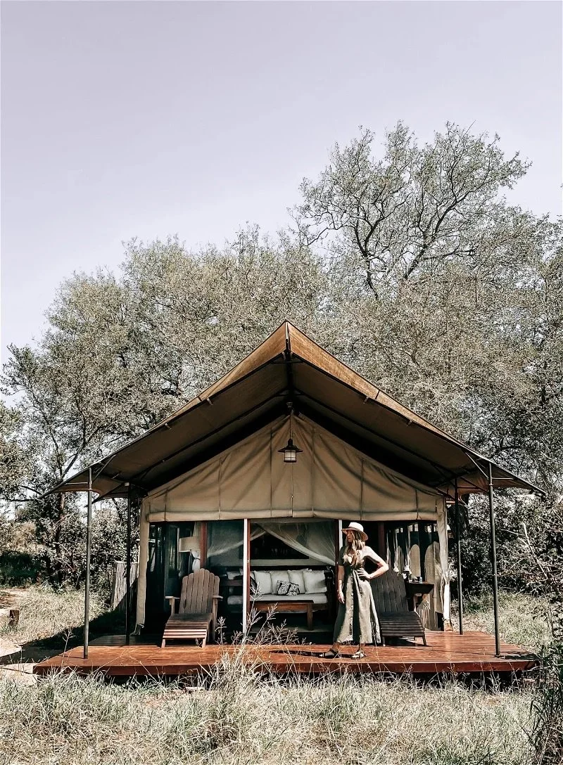 A woman standing on a wooden deck in front of a canvas glamping tent, surrounded by trees and grass.