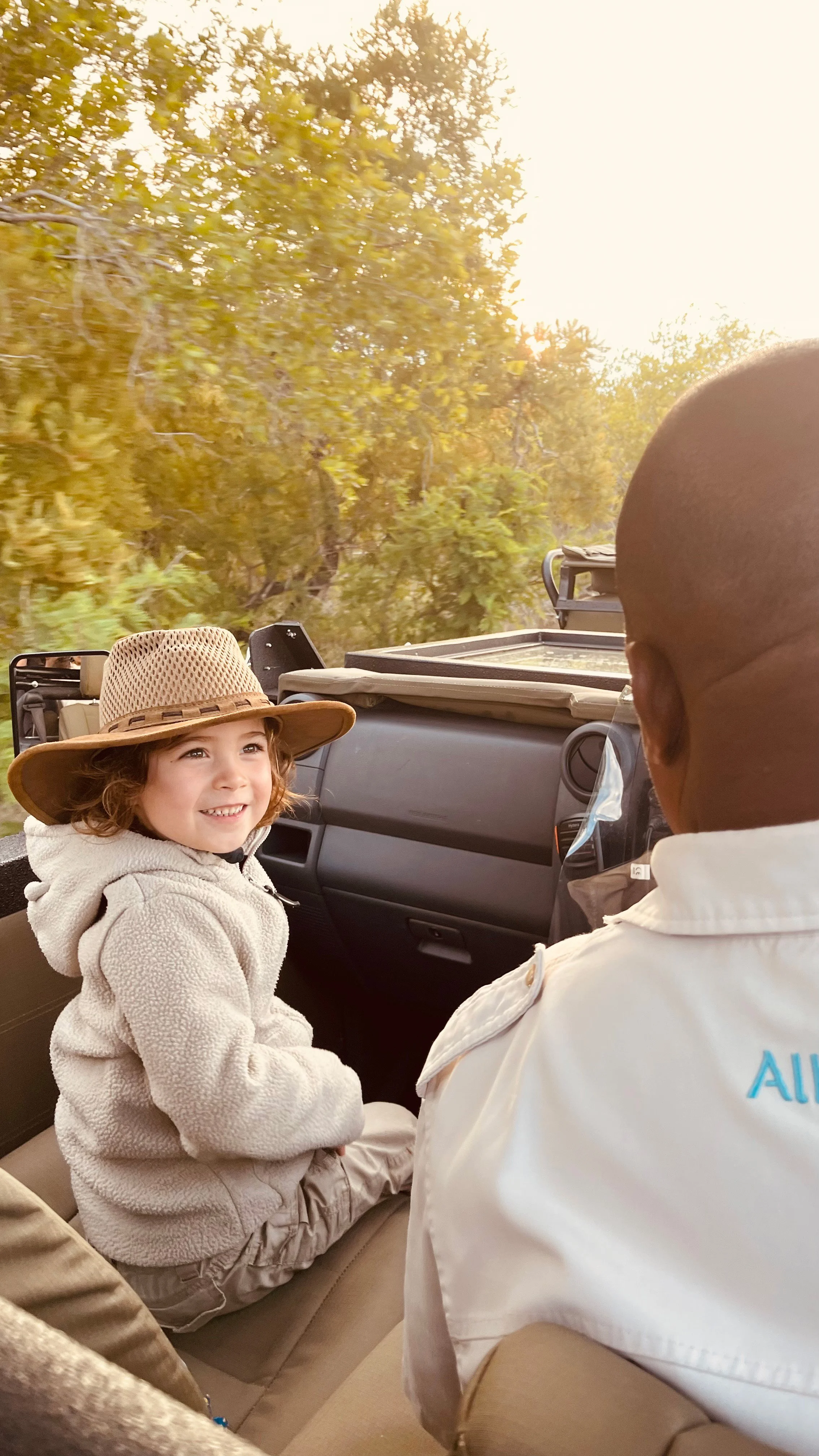 A young child with brown curly hair wearing a wide-brimmed straw hat and beige jacket, sitting in the front seat of a vehicle, smiling at an adult man facing away from the camera, with trees in the background.