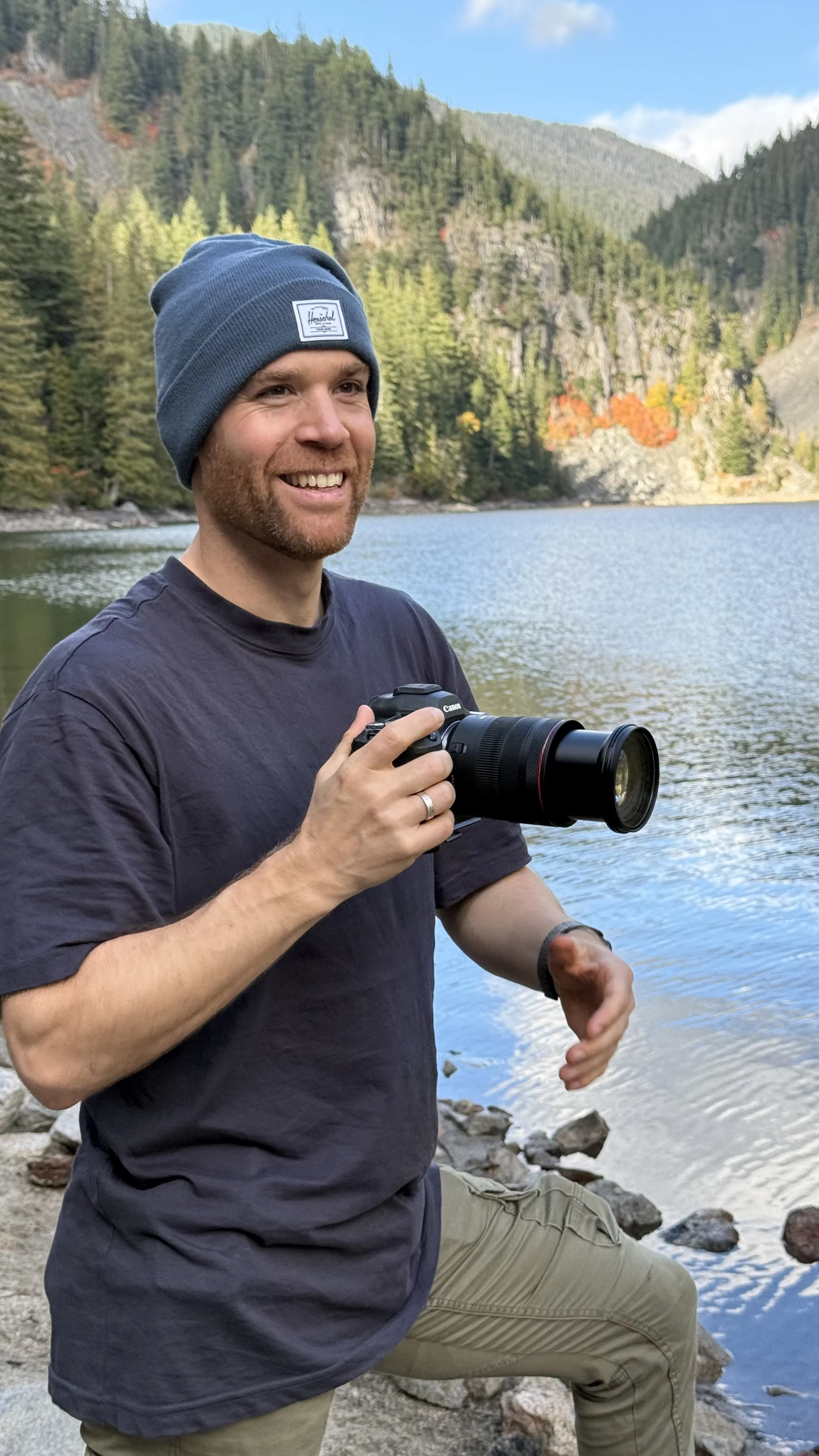 Erik van der Ven holding a camera outdoors near a lake with a backdrop of a forested mountainous landscape.