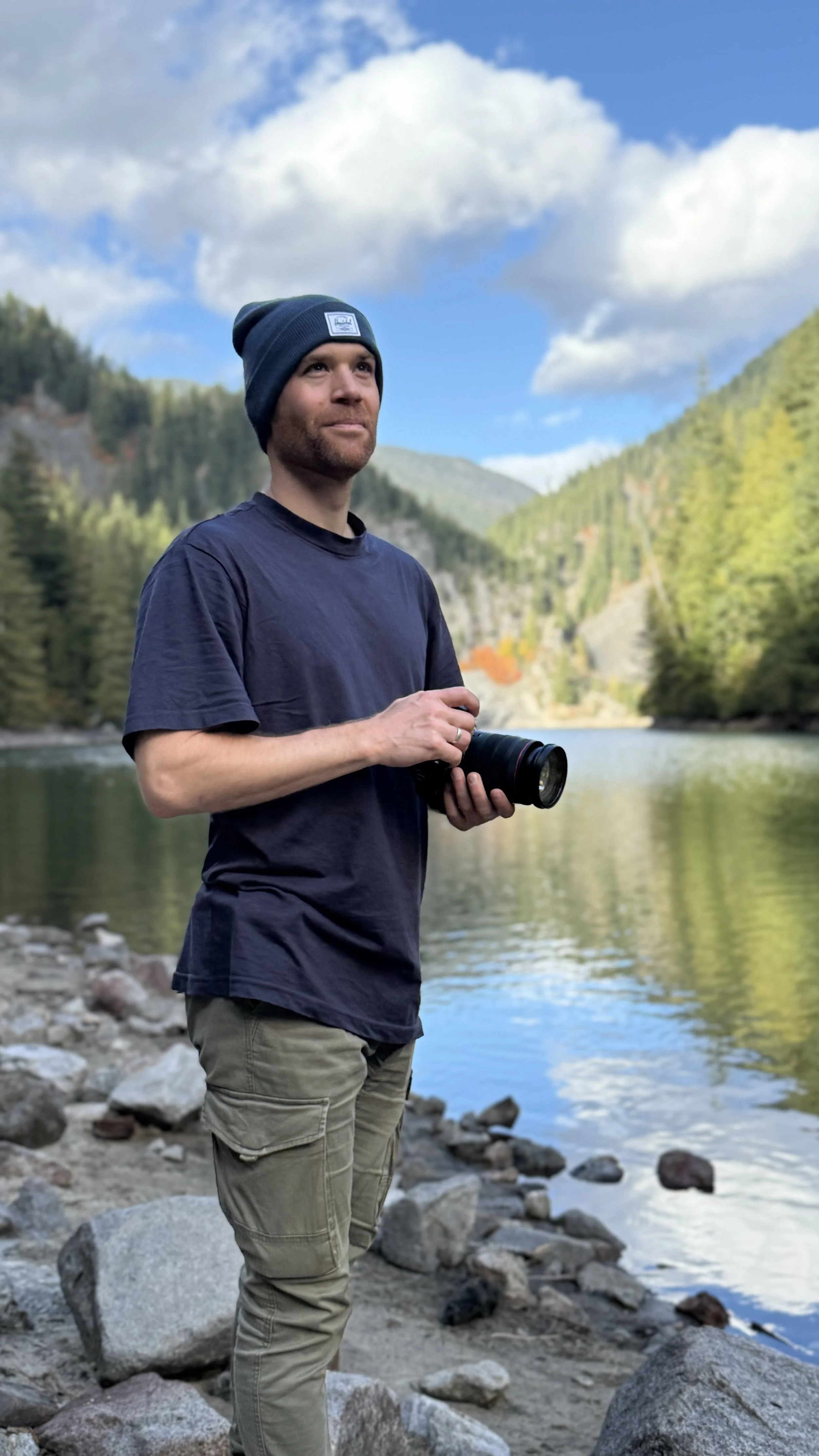 Erik van der Ven holding a camera, wearing dark t-shirt and beanie, with forested mountains and partly cloudy sky in background.