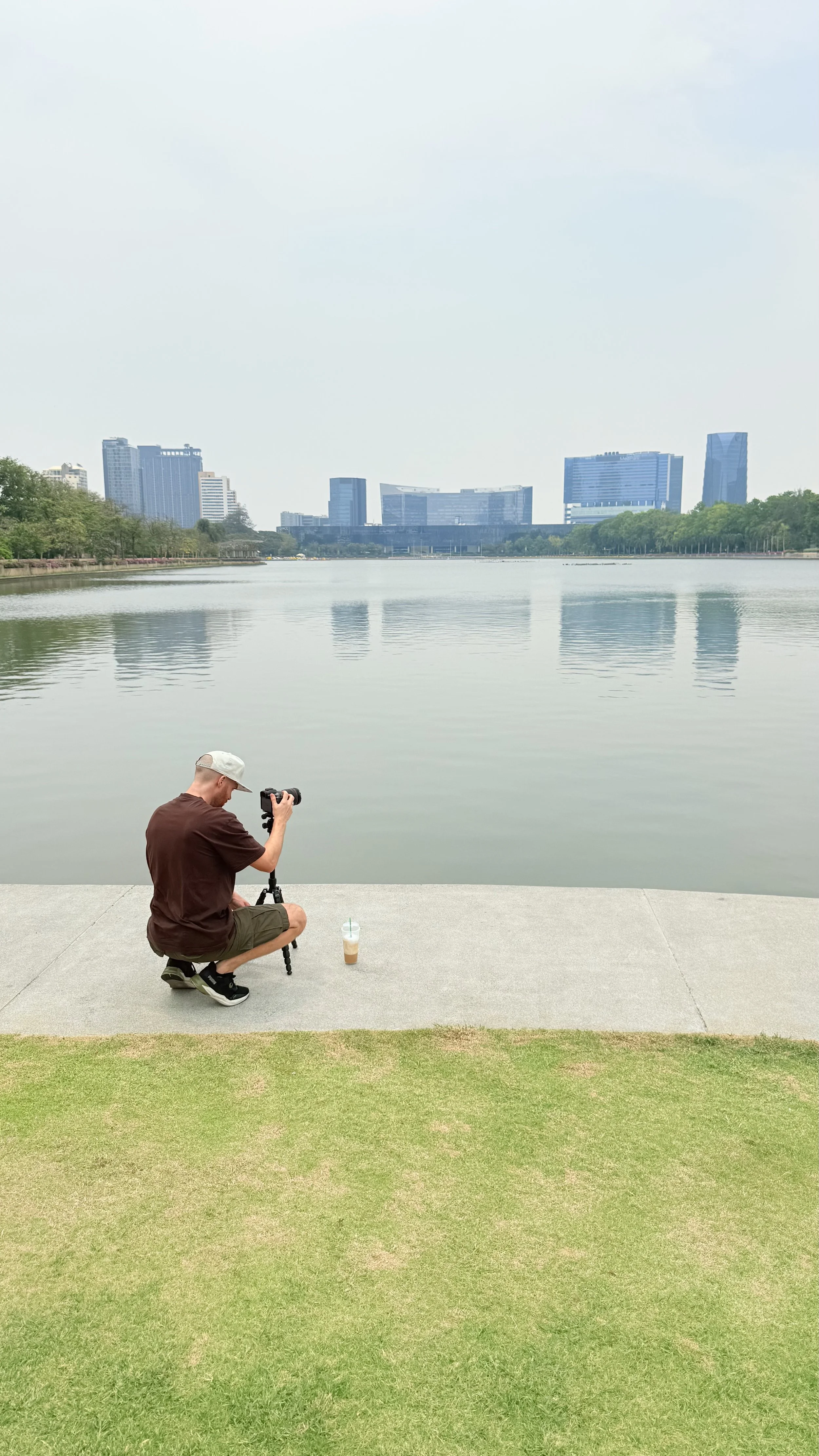 Erik van der Ven kneeling on a concrete walkway beside a lake takes a photograph with a camera on a tripod. There is a beverage in a plastic cup with a straw on the ground. The background shows calm water, a city skyline with tall buildings, and tree