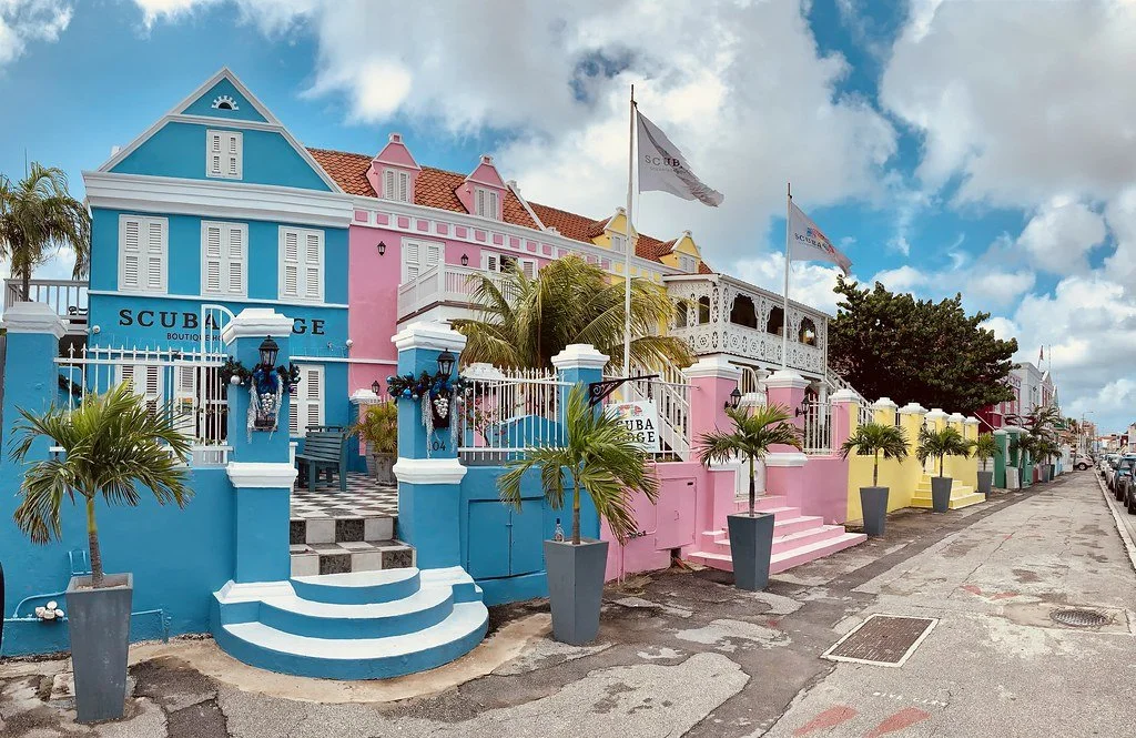 Colorful houses with pastel pink, blue, yellow, and green facades along a street, decorated with flags, potted palm trees, and white picket fences, under a partly cloudy sky.