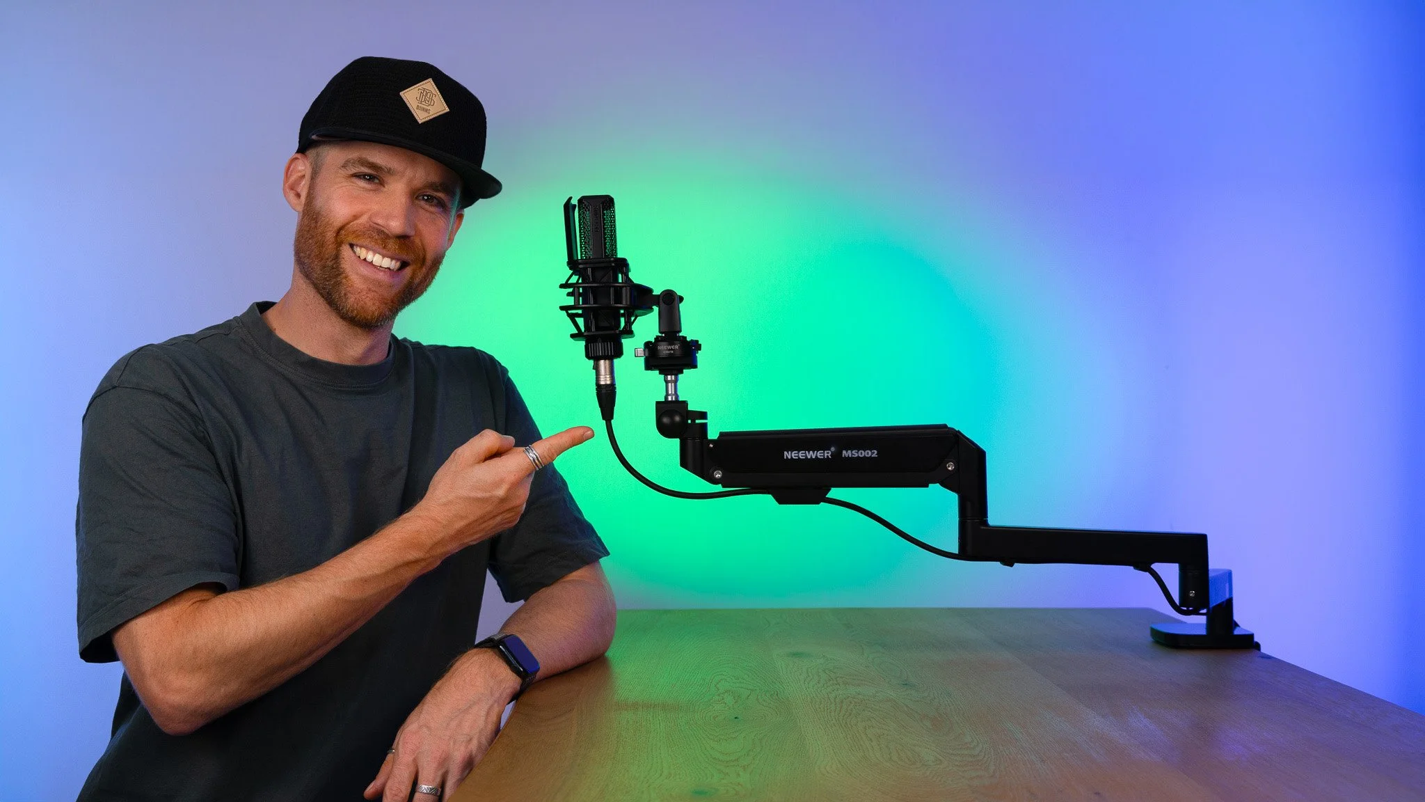 Erik van der Ven wearing a black cap and dark t-shirt point at a high-precision microphone arm a microphone, sitting at a wooden table with a colorful gradient background.