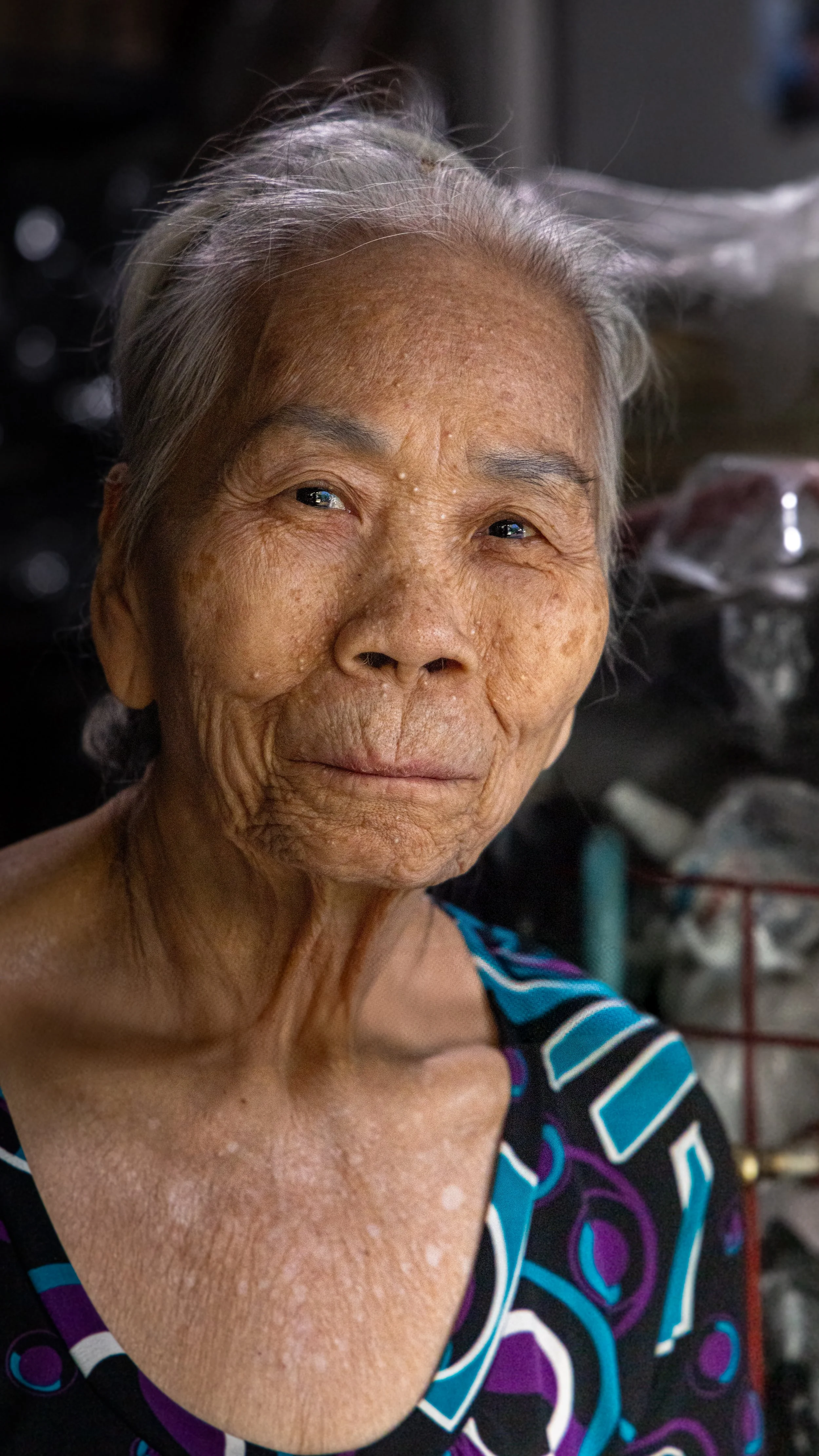 Close-up of an elderly woman with gray hair and a patterned shirt, smiling softly indoors.
