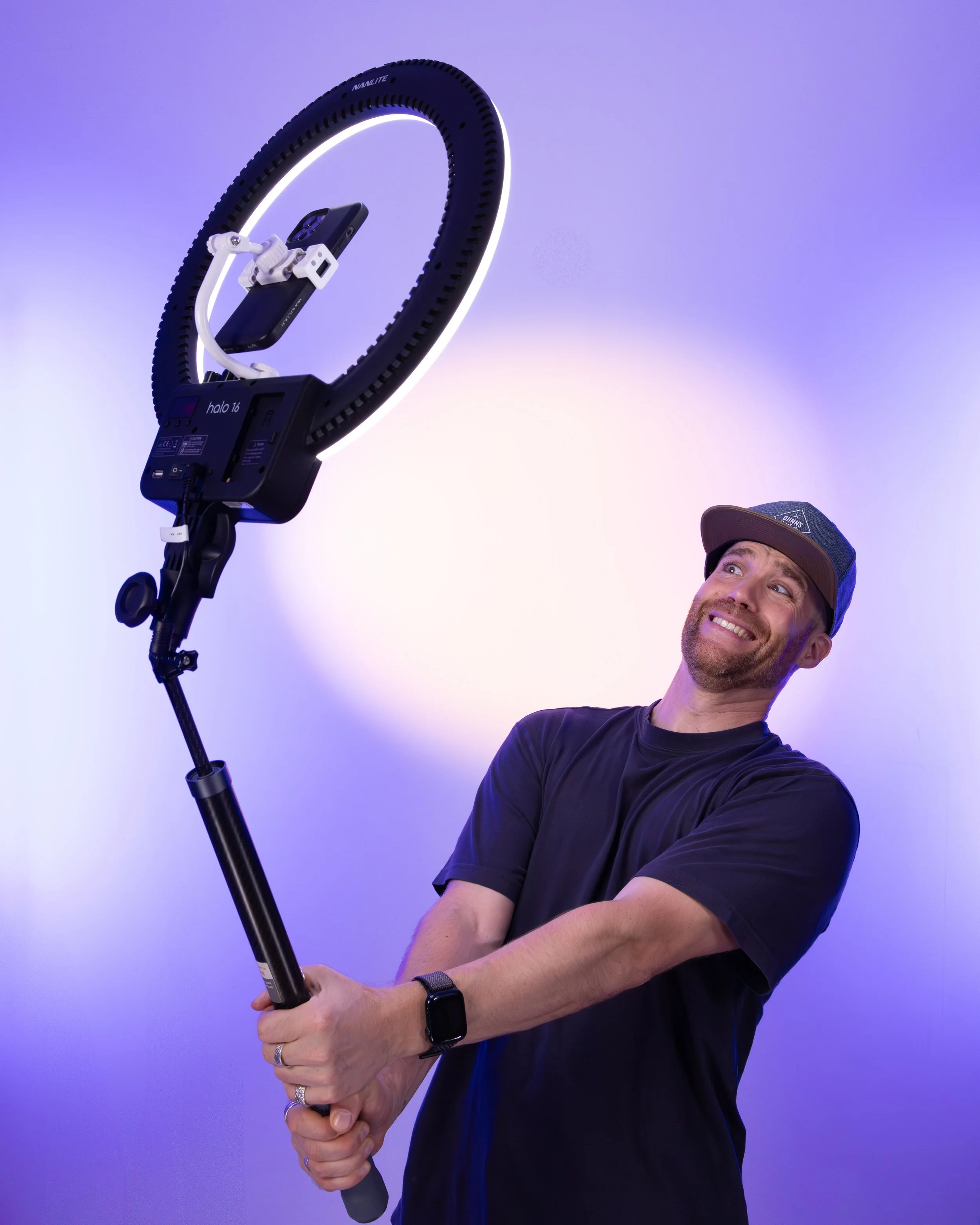 Erik van der Ven smiling while holding a selfie stick with a mounted smartphone, wearing a cap and a black shirt, against a gradient purple and blue background.