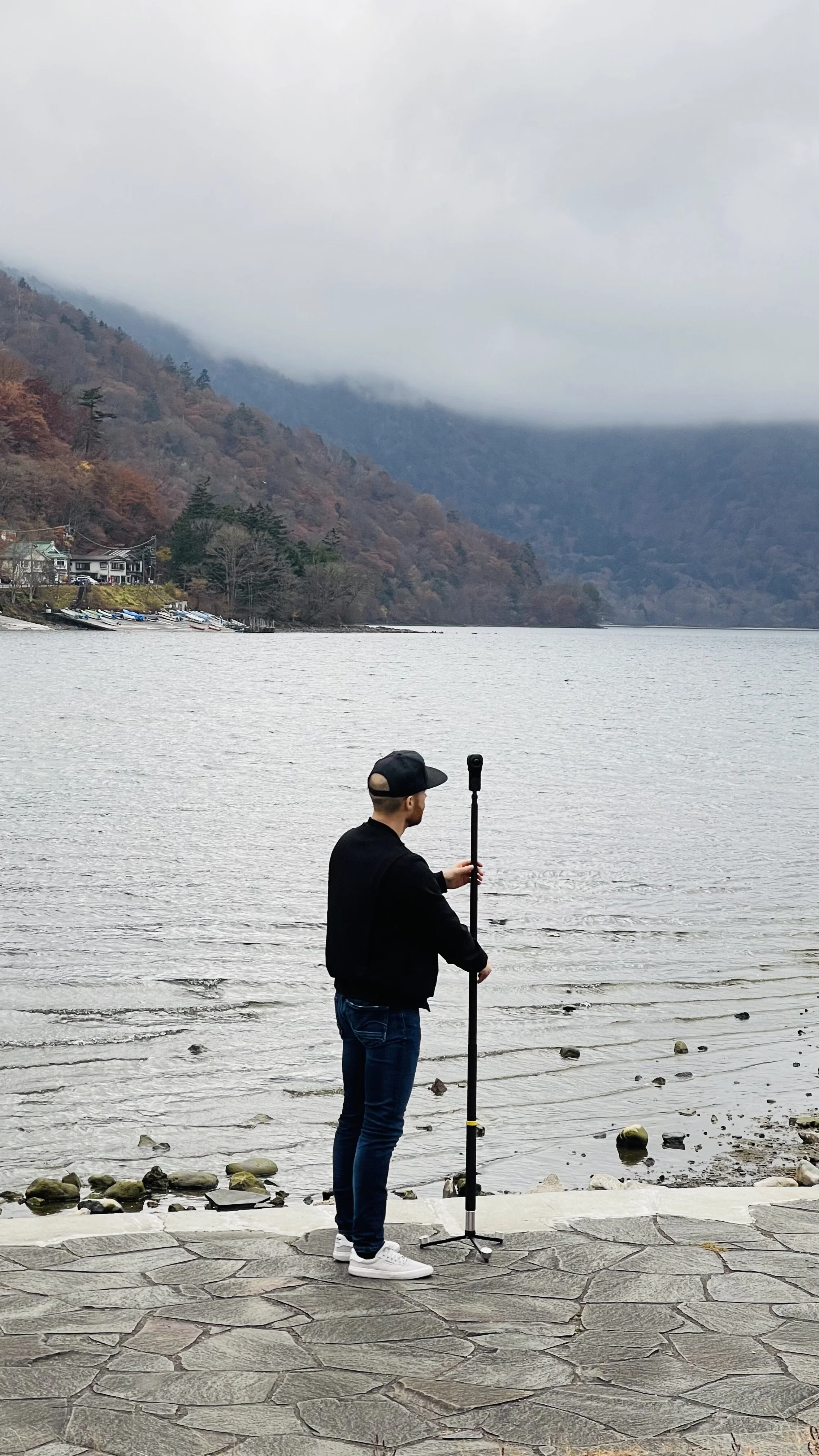 Erik van der Ven wearing a black cap, black jacket, and jeans stands at the edge of a lake with rocky shore, holding a selfie stick. In the background, there are leafless trees, hills covered in autumn-colored foliage, a cloudy sky, and a mountainous