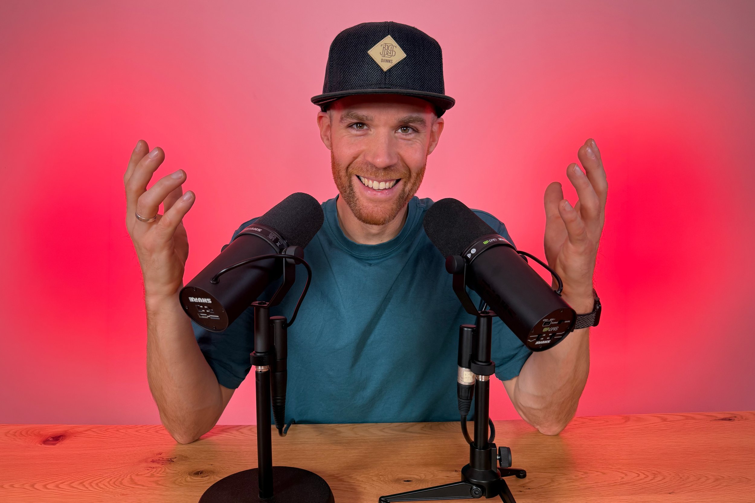 Erik van der Ven with a beard and blue t-shirt sitting at a wooden table with two microphones in front of him in front of a red gradient background. He is smiling and wearing a black cap with a beige logo, and is holding his hands up near his face.
