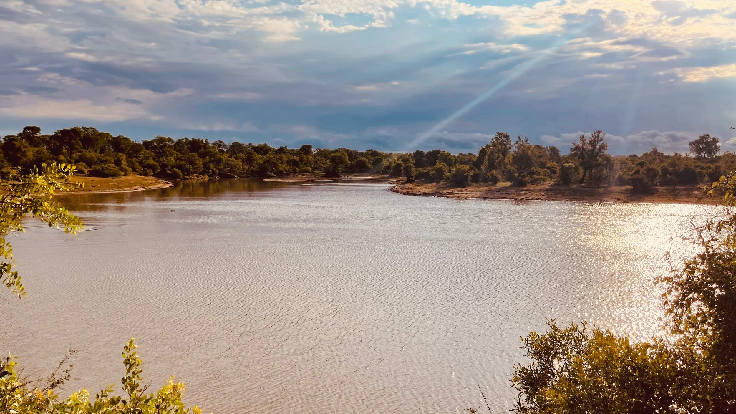 A serene river landscape with calm water reflecting the sky, surrounded by green trees and bushes, under a partly cloudy sky with sunlight.