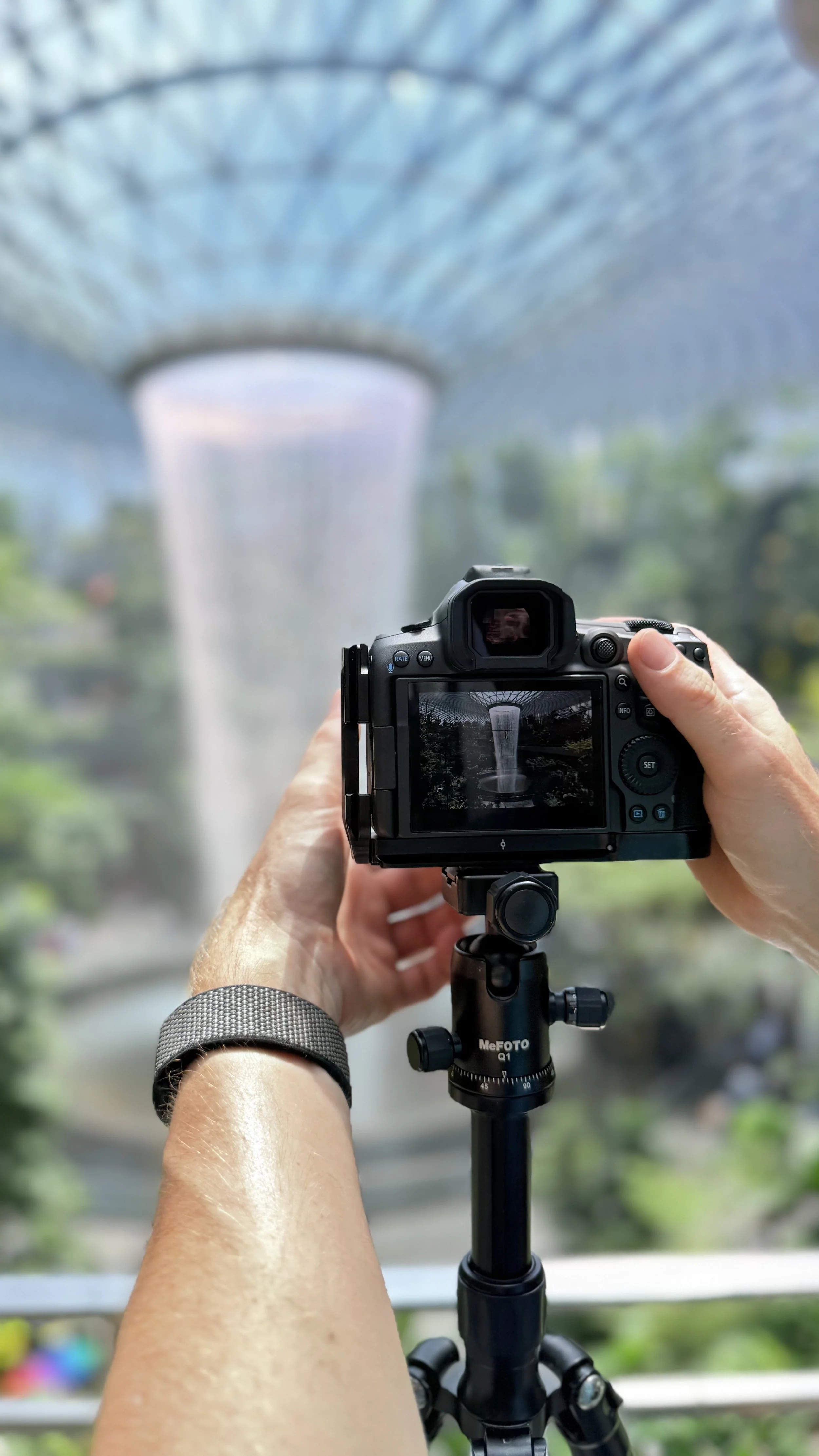 Erik van der Ven taking a photograph the Rain Vortex in Jewel Changi Airport Singapore, viewed through a camera on a tripod, during daytime.