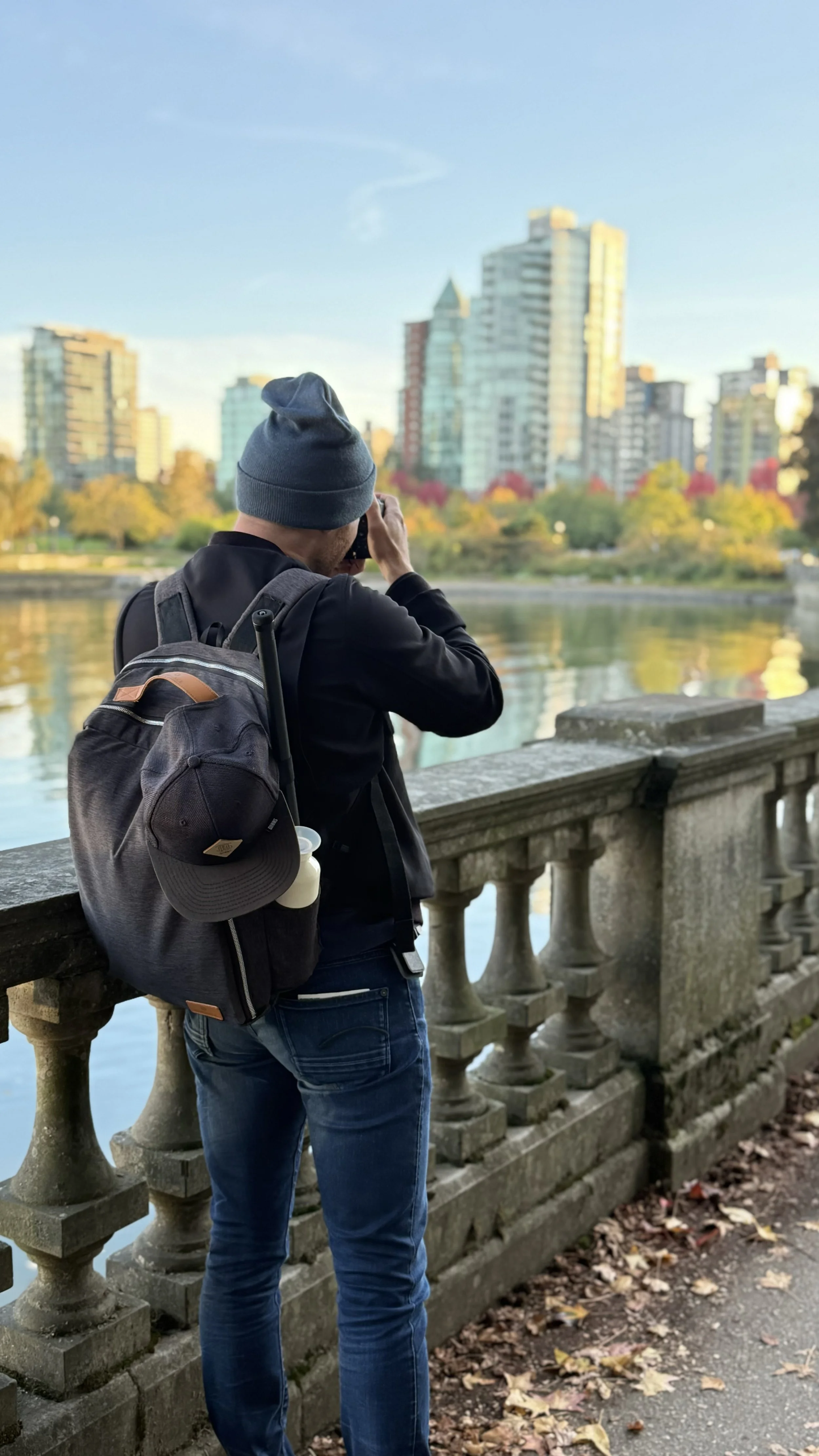 Erik van der Ven wearing a blue beanie and black jacket standing by a stone railing, taking a picture of the Vancouver city skyline across a body of water in the late afternoon or early evening, with fall foliage on the trees.