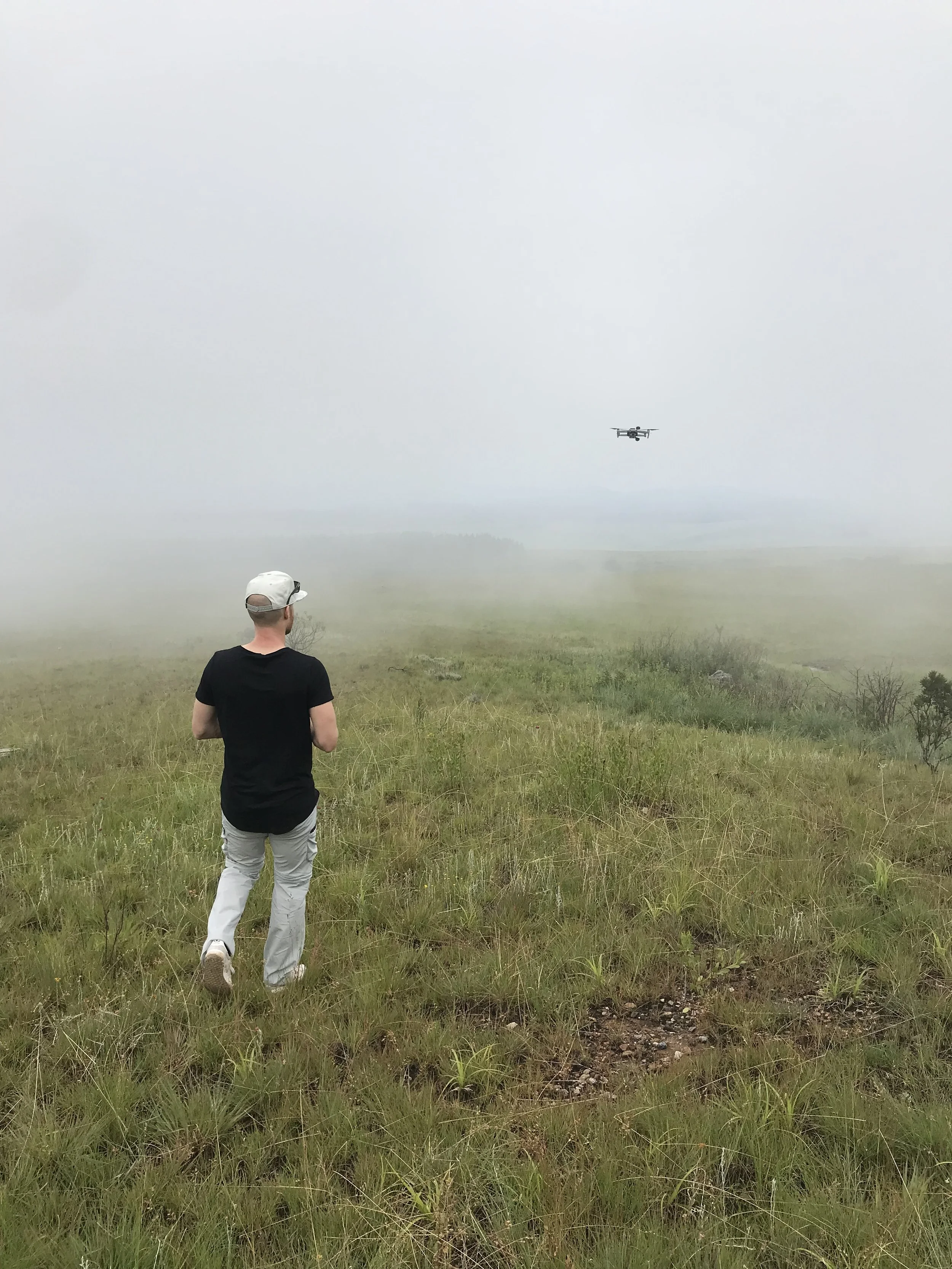 Erik van der Ven wearing a black shirt, light-colored pants, and a white cap is walking in a grassy field with tall grass and small bushes. In the background, a drone is flying in the cloudy sky above the field.
