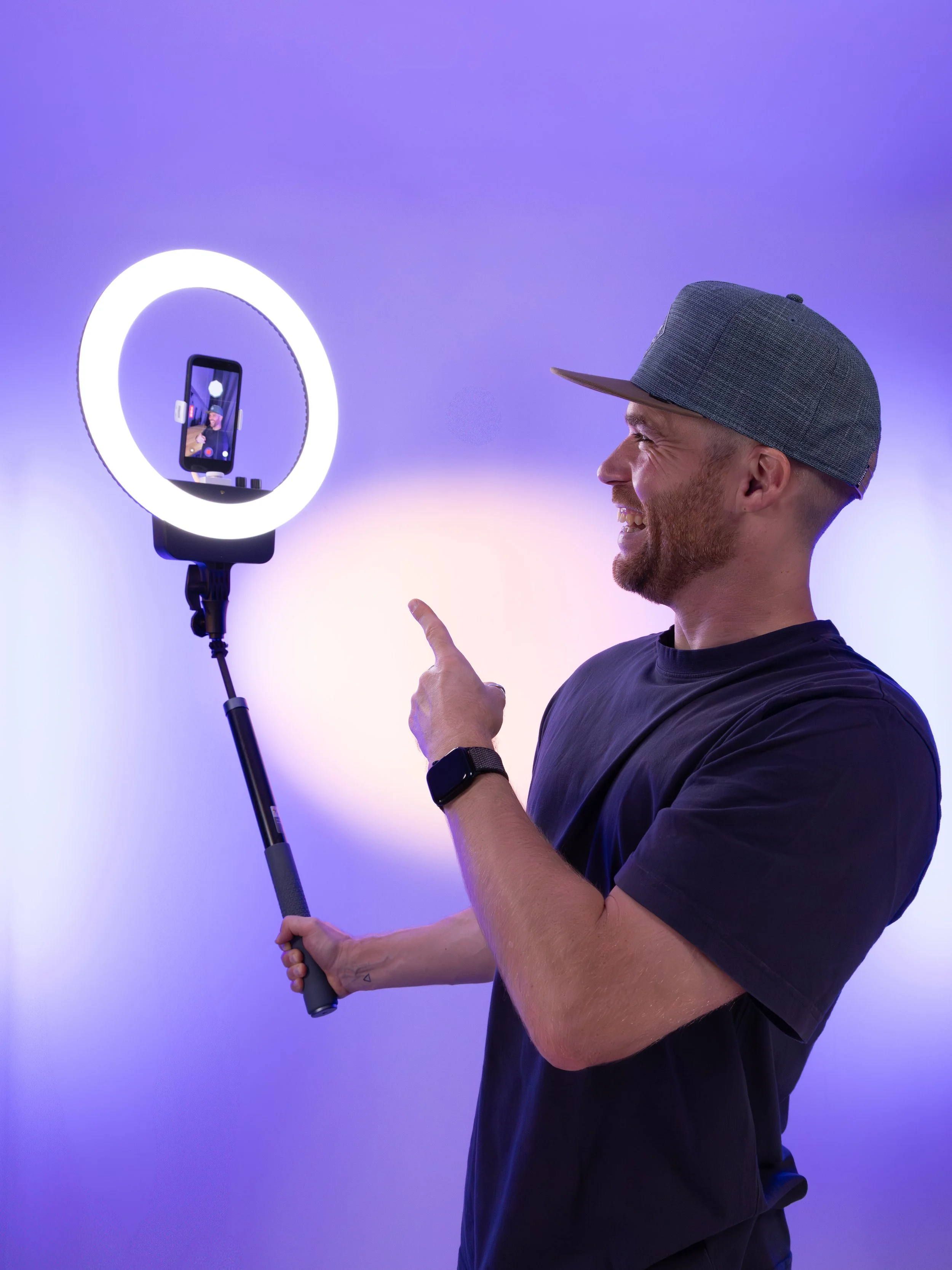 Erik van der Ven waring a black T-shirt, a gray baseball cap, and a watch, is smiling and pointing with his index finger at a smartphone mounted on a selfie stick. The smartphone screen shows Erik and a bright ring light behind him.