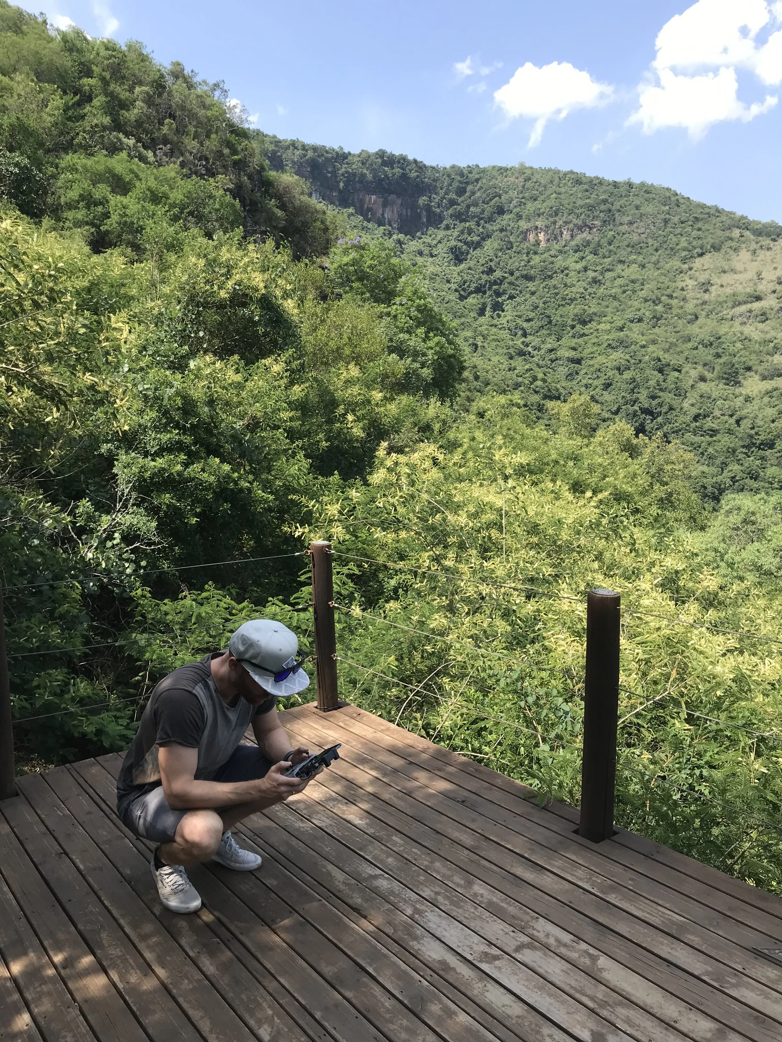 Erik van der Ven crouching on wooden deck with lush green trees and a mountain in the background, holding a device in his hand, under a partly cloudy sky.
