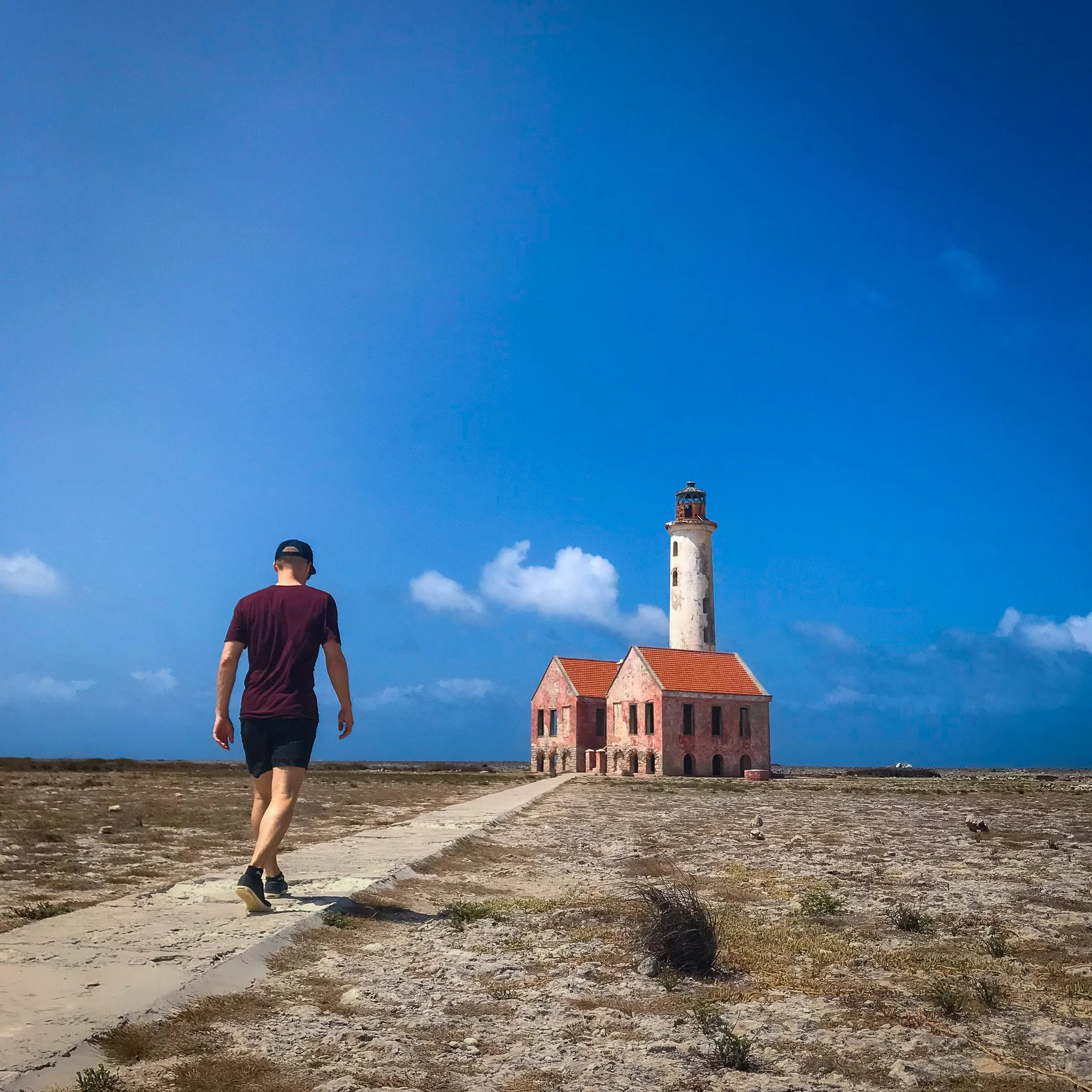 A person wearing a maroon shirt and black shorts walking down a concrete path on a barren landscape with patches of grass. In the background, there is a lighthouse with a red roof and a small building with a red tiled roof. The sky is blue with scattered white clouds.