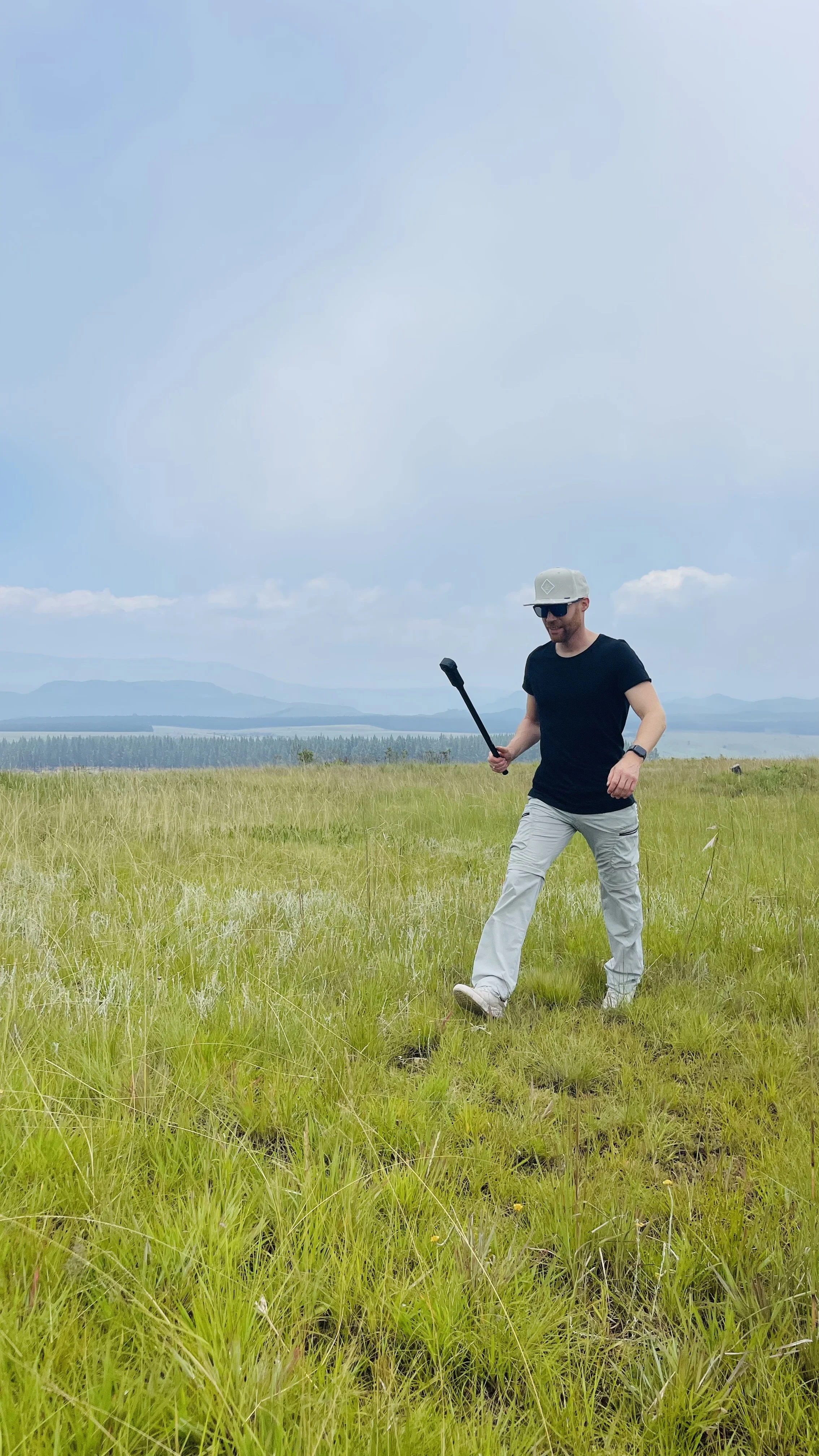Erik van der Ven wearing a black t-shirt, light gray pants, a white hat, and sunglasses walks through a grassy field holding a selfie stick. The background shows a blue sky with some clouds and distant mountains.
