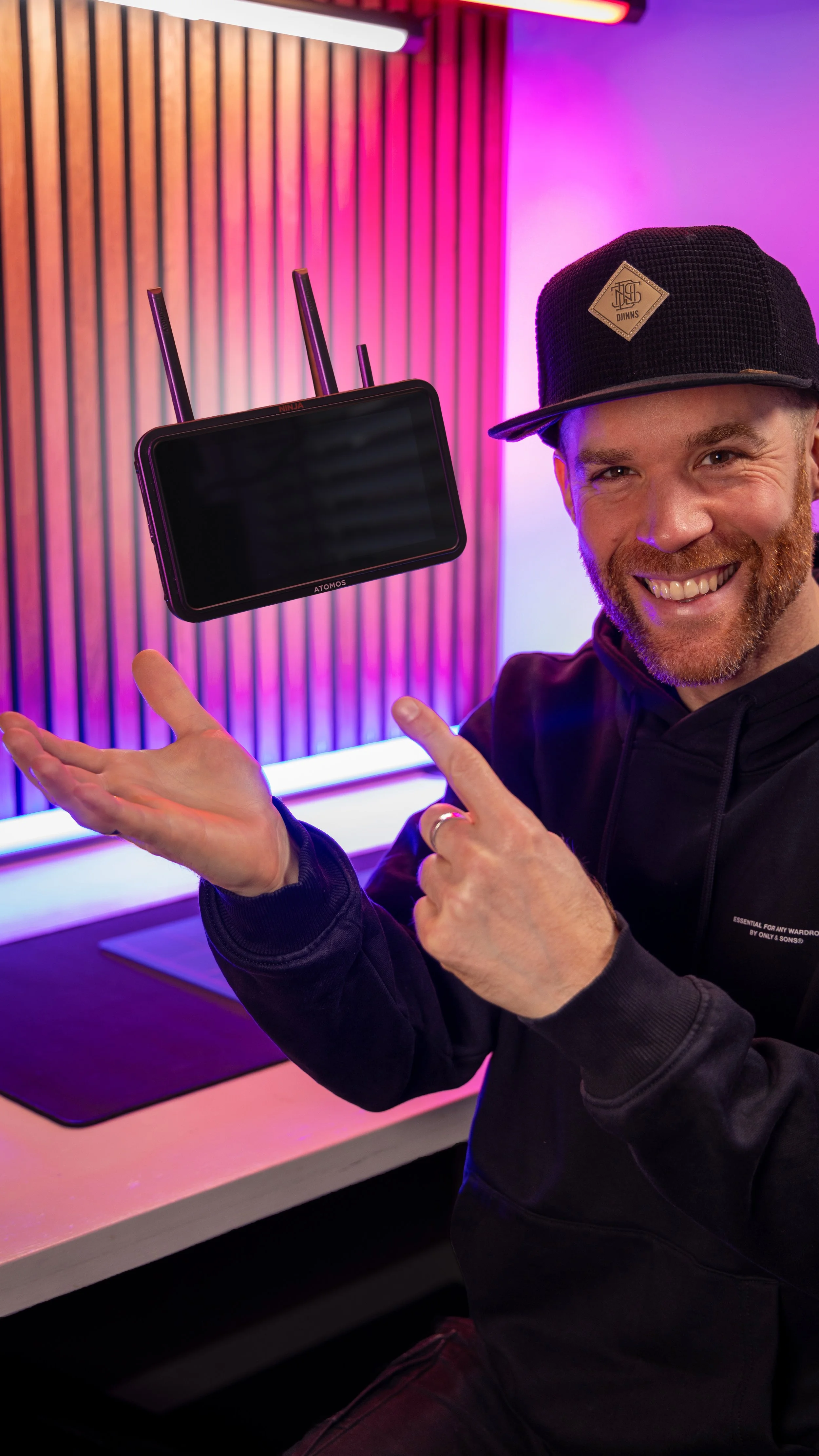 Erik van der Ven with a beard and hat smiling and pointing at a screen with antennas. The background features colorful neon lighting and a wooden wall.
