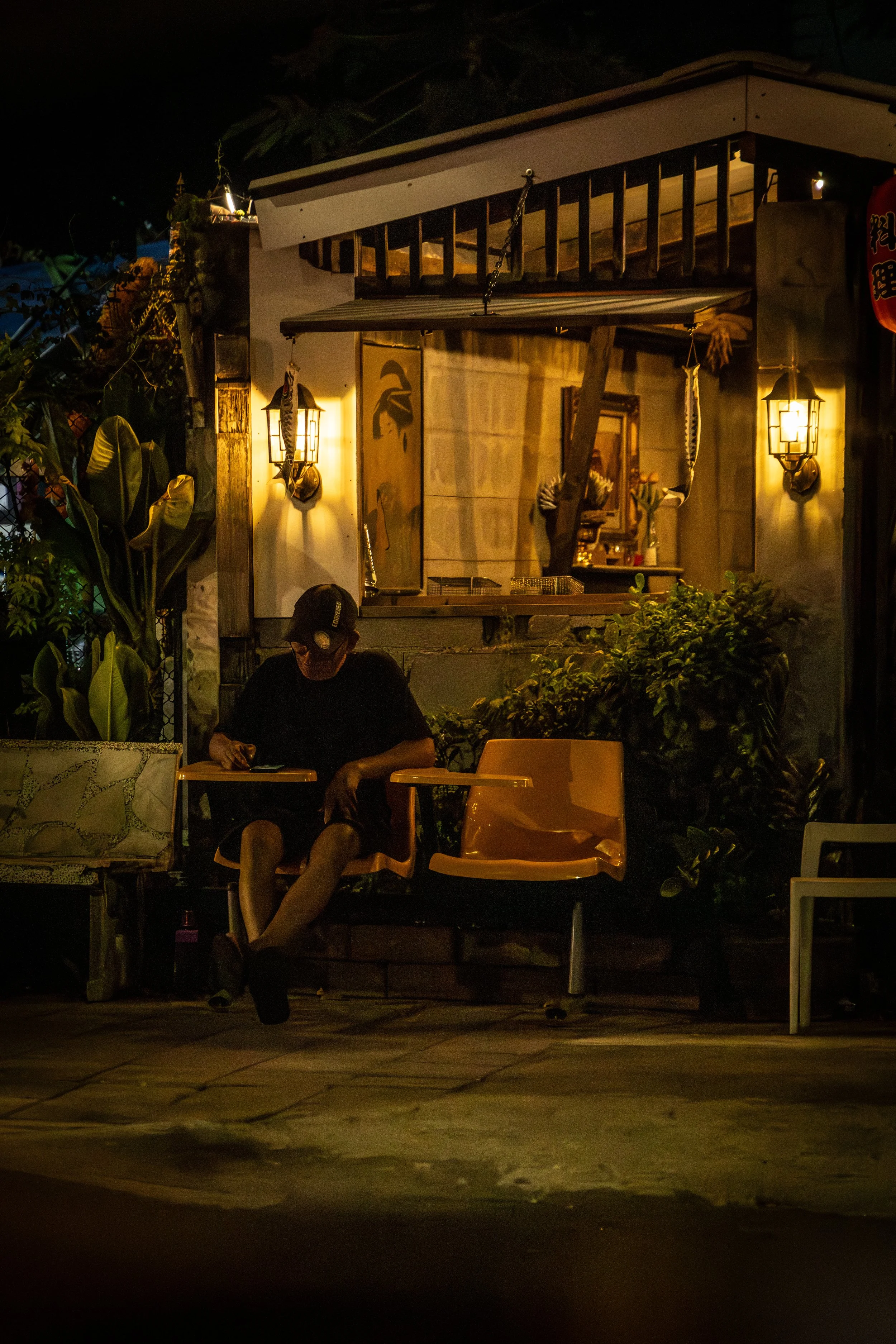 Nighttime scene showing a man sitting on a bench outside a lit-stained building, with plants and lanterns around.