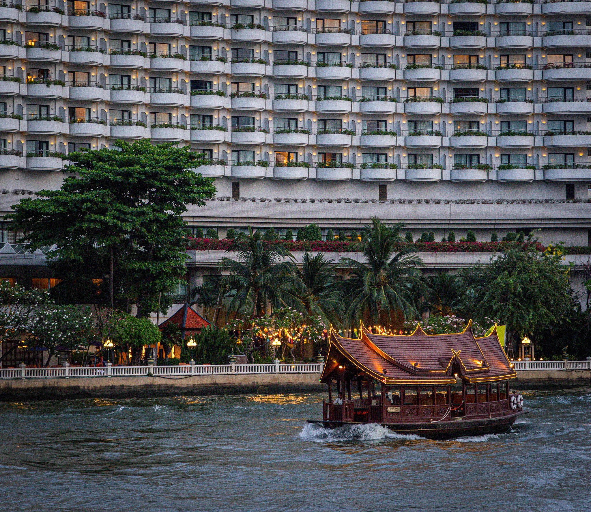 A large hotel with multiple balconies illuminated at dusk, situated along a river with tropical trees and plants in the foreground. A traditional-style boat with a roof and lights is on the water with passengers on board.