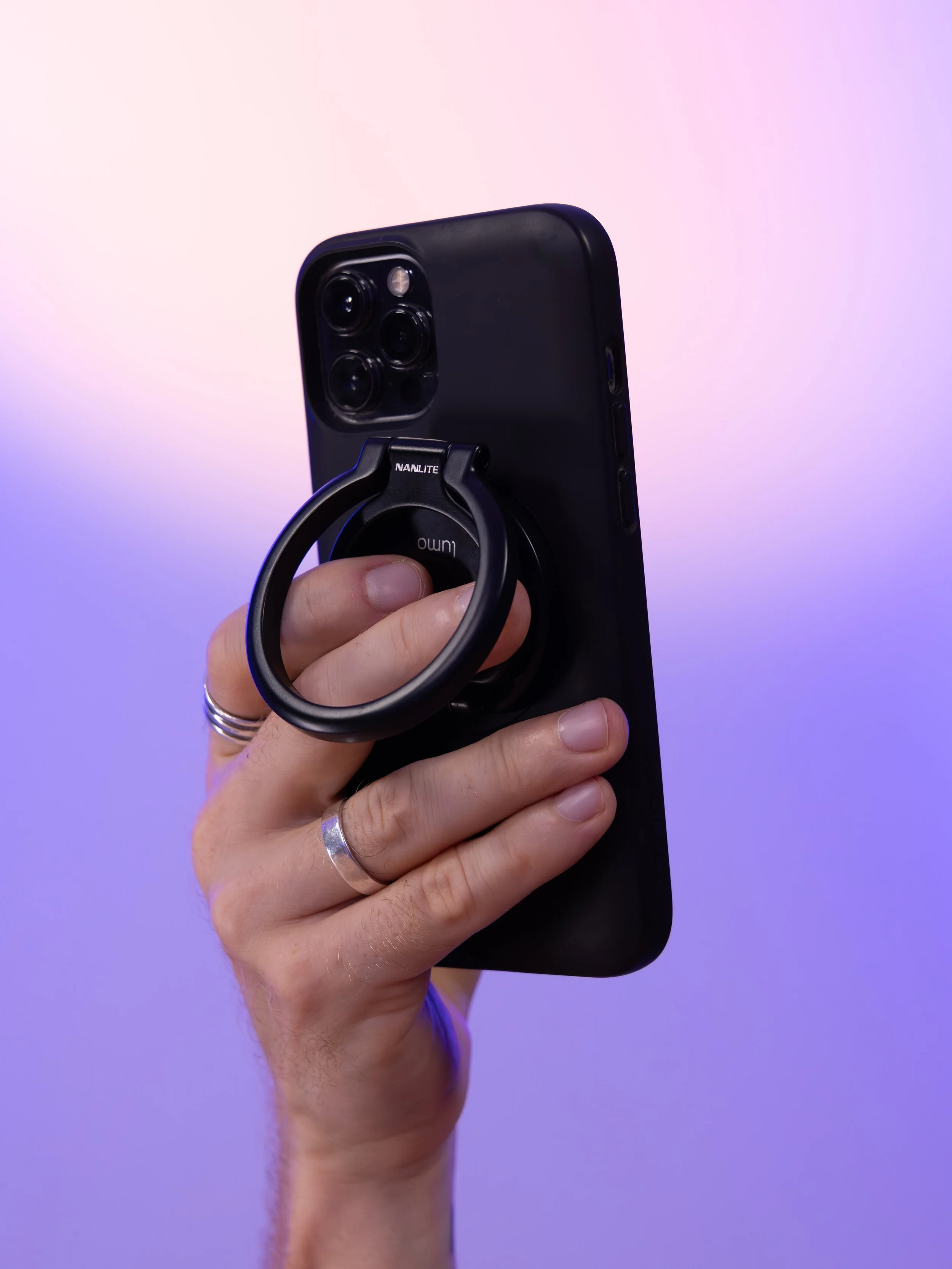 A hand holding a black smartphone with a ring holder attached, against a gradient background of soft purple and pink hues.